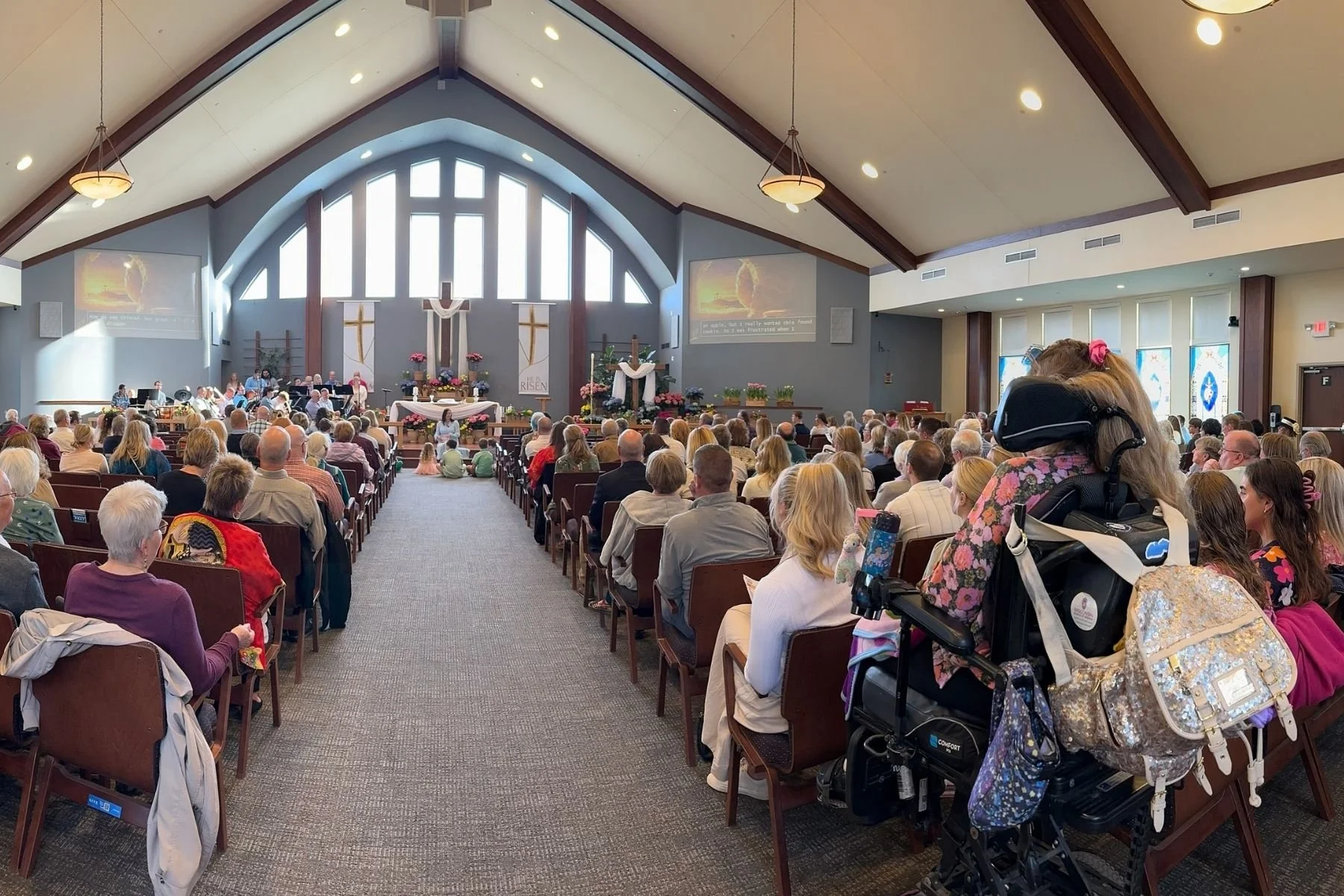 A congregation inside a church during a service, with people seated in pews facing the altar. The church features high, arched windows, a cross, and religious banners at the front. Some people, including a woman with a decorated backpack and a stroller, are visible in the foreground.