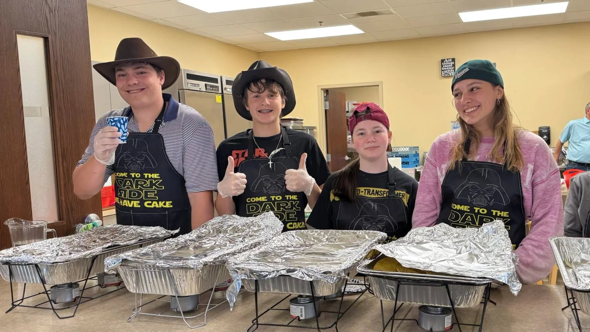 Four teenagers wearing Star Wars-themed aprons with Darth Vader's face and the words "Come to the Dark Side, We Have Cake" stand behind a table covered with foil-covered trays of food. They are smiling, and some are giving thumbs up.