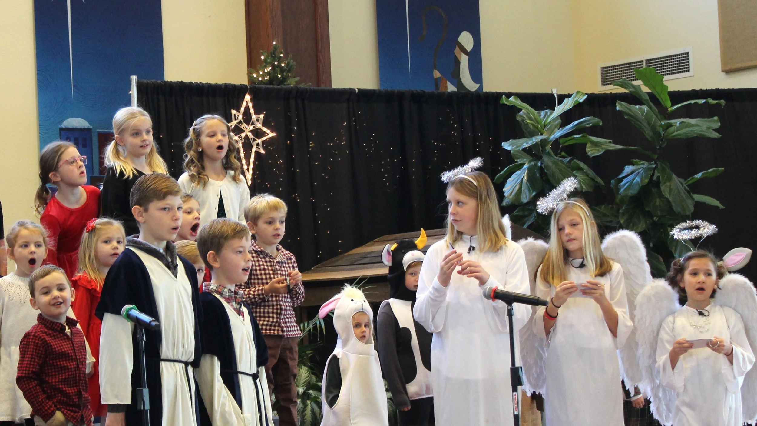 Children's choir performing a holiday concert on stage with Christmas decorations, including a star and a small decorated Christmas tree.