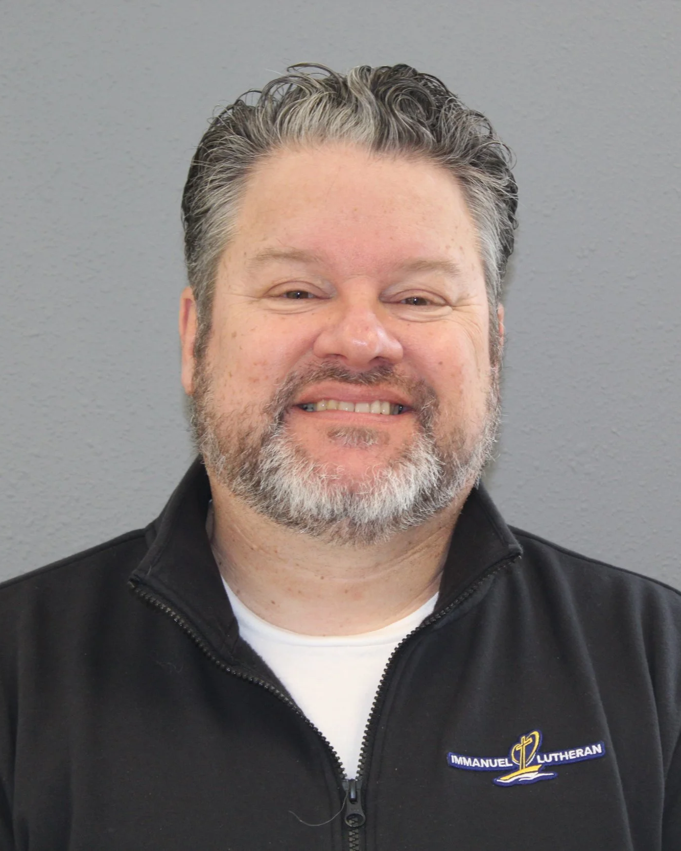 A smiling middle-aged man with gray and black hair and a beard, wearing a black zip-up jacket with an embroidered logo that says 'Immanuel Lutheran' and features a cross and a boat on water.