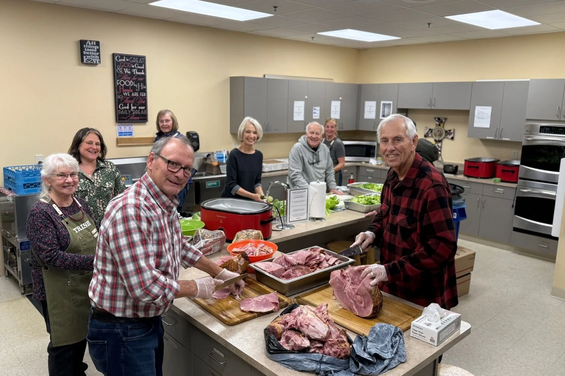 A group of people in a kitchen preparing meat for cooking. They are smiling and working together, with various cuts of meat on the counter and some leafy greens nearby.