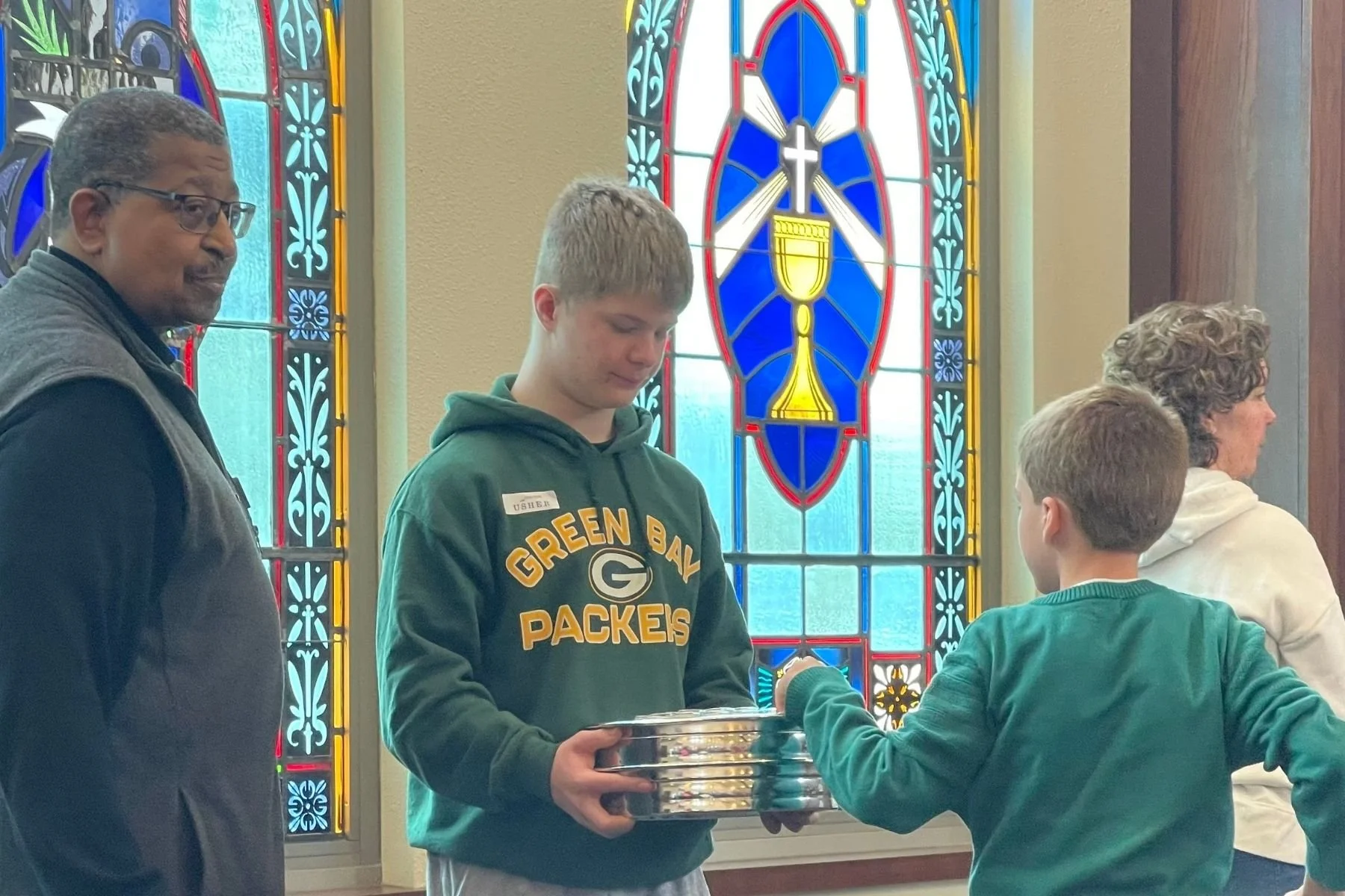 A young man with short blonde hair, wearing a Green Bay Packers hoodie, holds a silver tray while a young boy in a green jacket receives something from him in a church. An older woman with curly gray hair stands behind the boy, and another man with short dark hair and glasses, dressed in dark clothing, looks towards the young man. The background features stained glass windows with religious symbols.