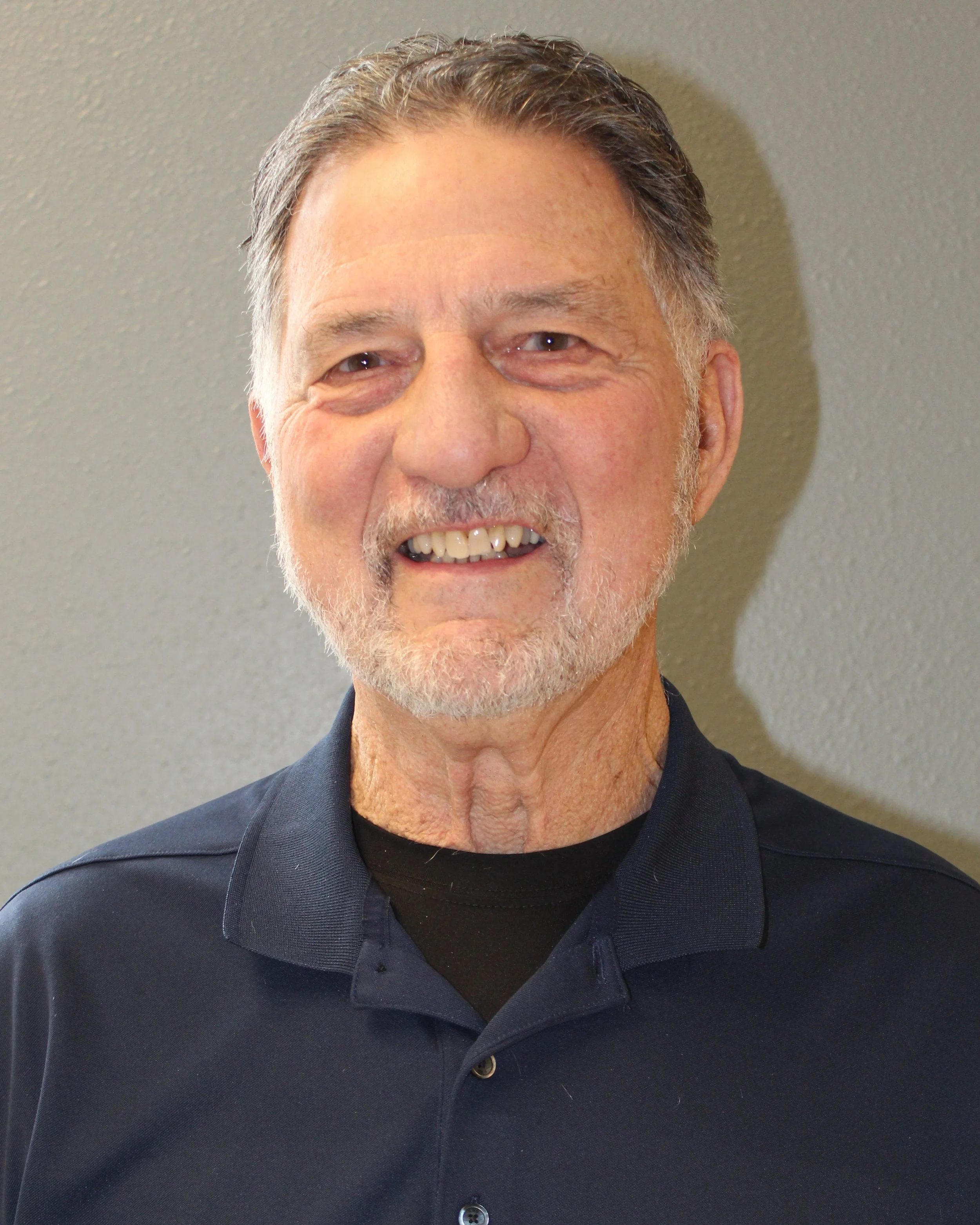 Close-up of a smiling older man with gray hair and a beard, wearing a dark blue collared shirt, standing against a plain beige wall.
