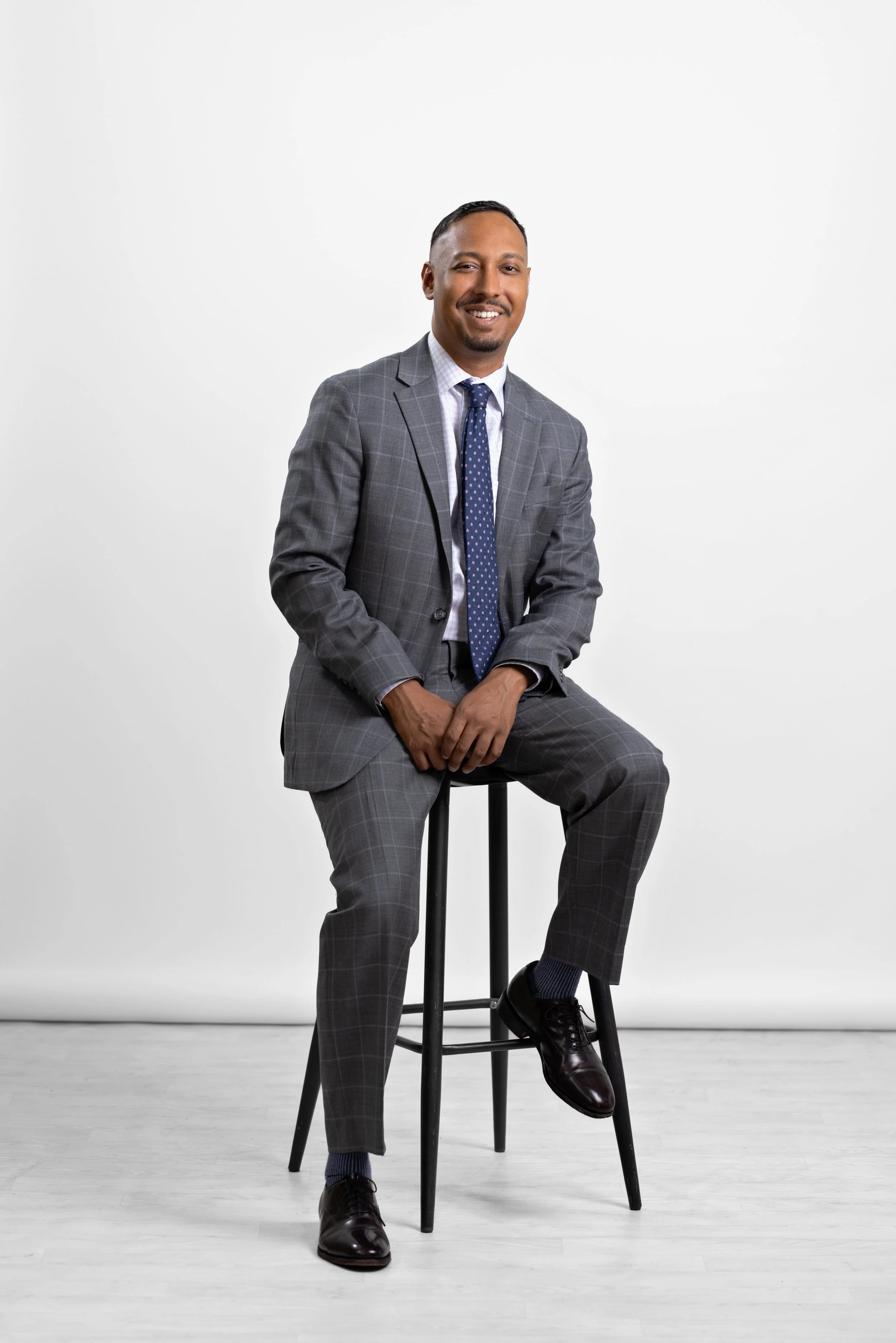 A man in a gray checked suit sitting on a black stool against a white background, smiling, with a blue polka dot tie and black dress shoes.