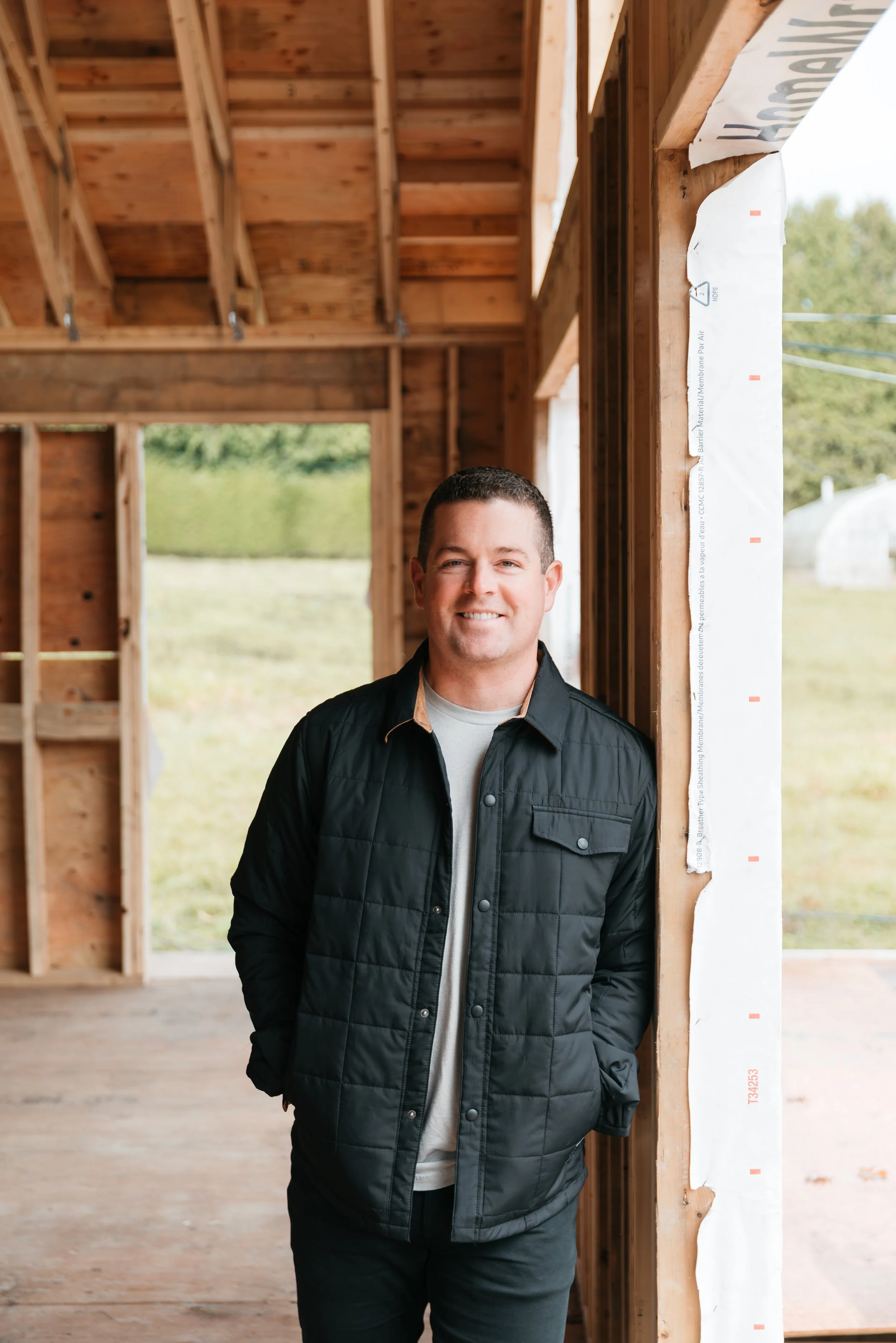 A man standing inside a wooden house under construction, smiling at the camera, with a background of open windows and green outdoor scenery.