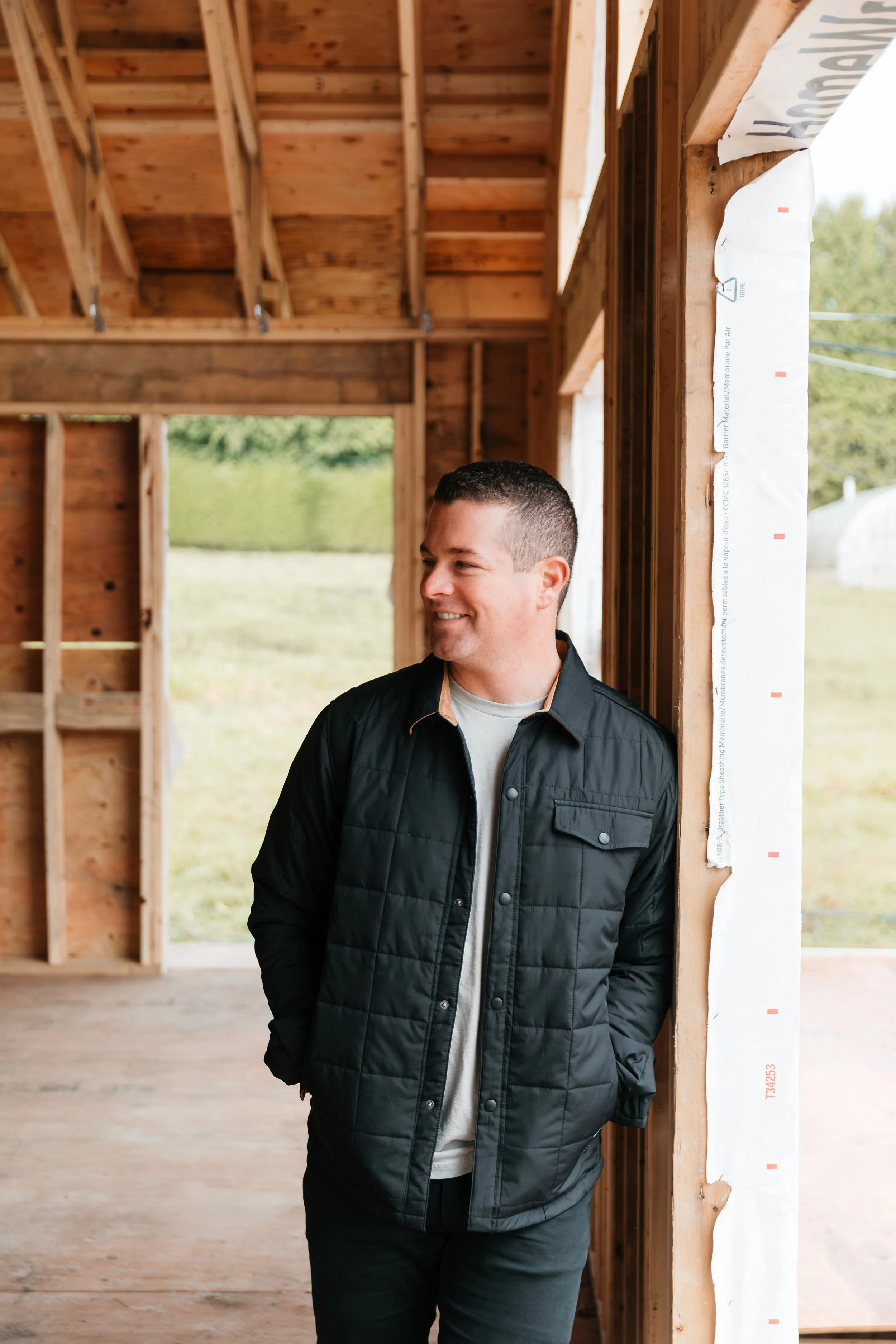 A man stands inside an unfinished wooden house, leaning against a window frame and looking away while smiling.