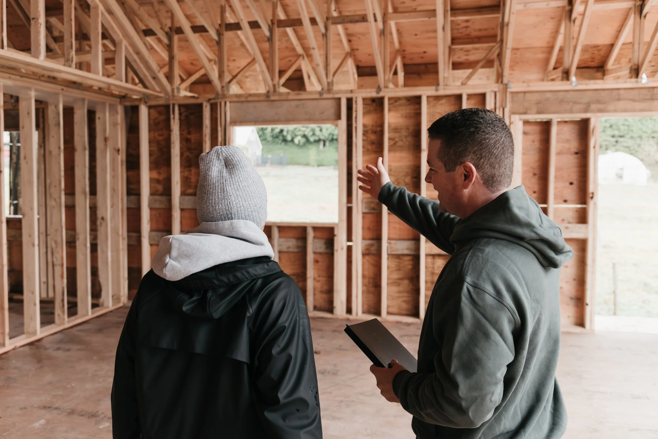 Two men examining the interior of a wooden house under construction, with exposed wooden framing and a large window showing an outdoor view.
