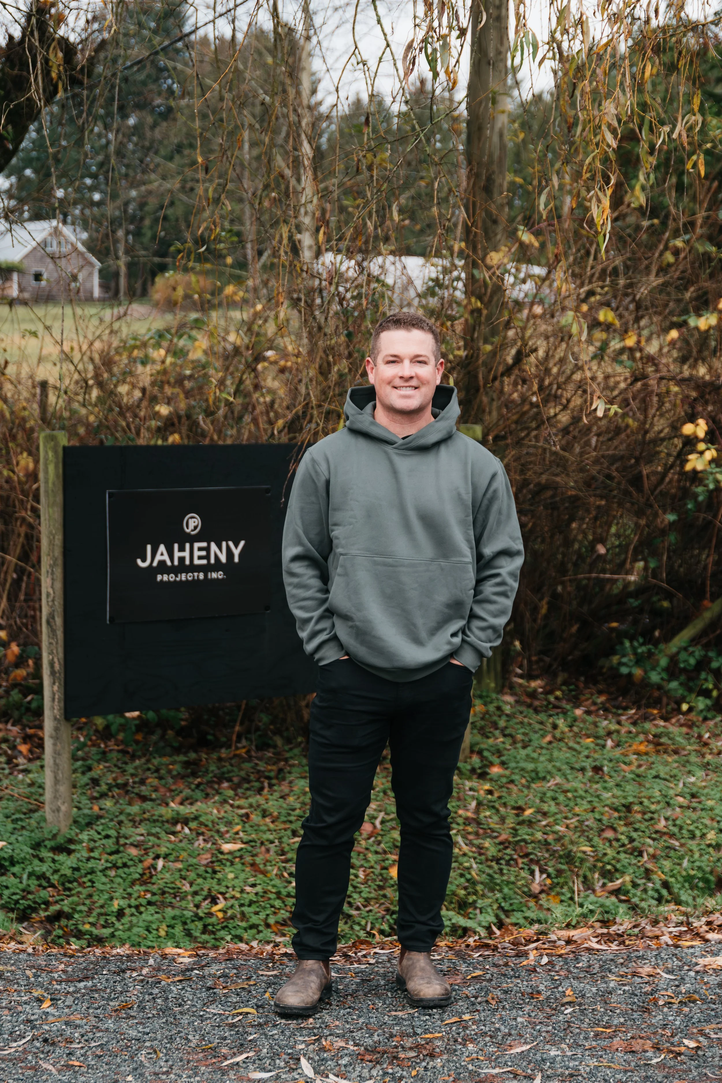 A man stands outdoors on a gravel path, smiling, with hands in his hoodie pockets. He is wearing a gray hoodie, black pants, and brown boots. Behind him is dense, leafless shrubbery and trees, with a black sign that reads 'JAHENY PROJECTS INC.'.