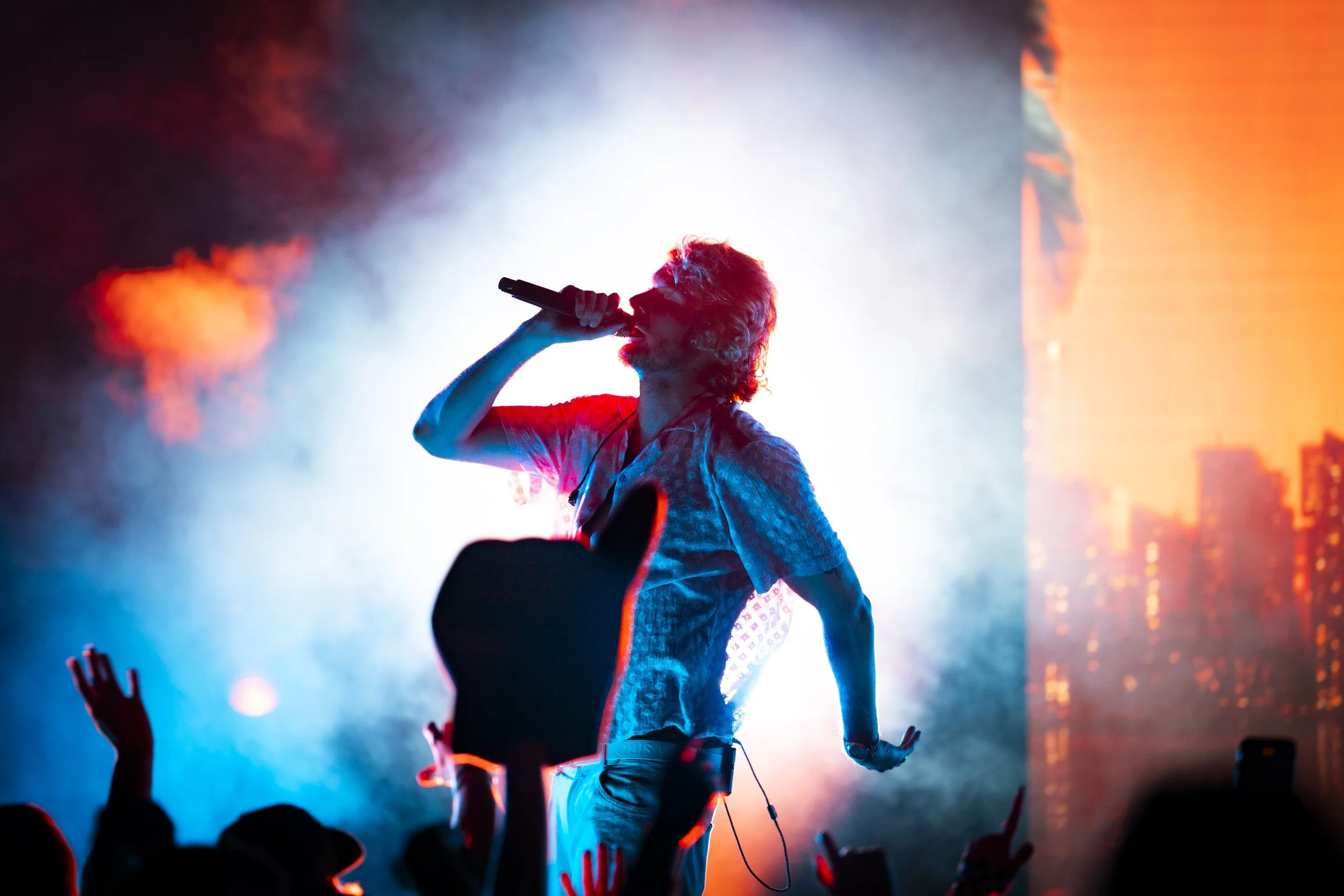 A young man with curly hair playing an electric guitar and singing into a microphone during a live performance.