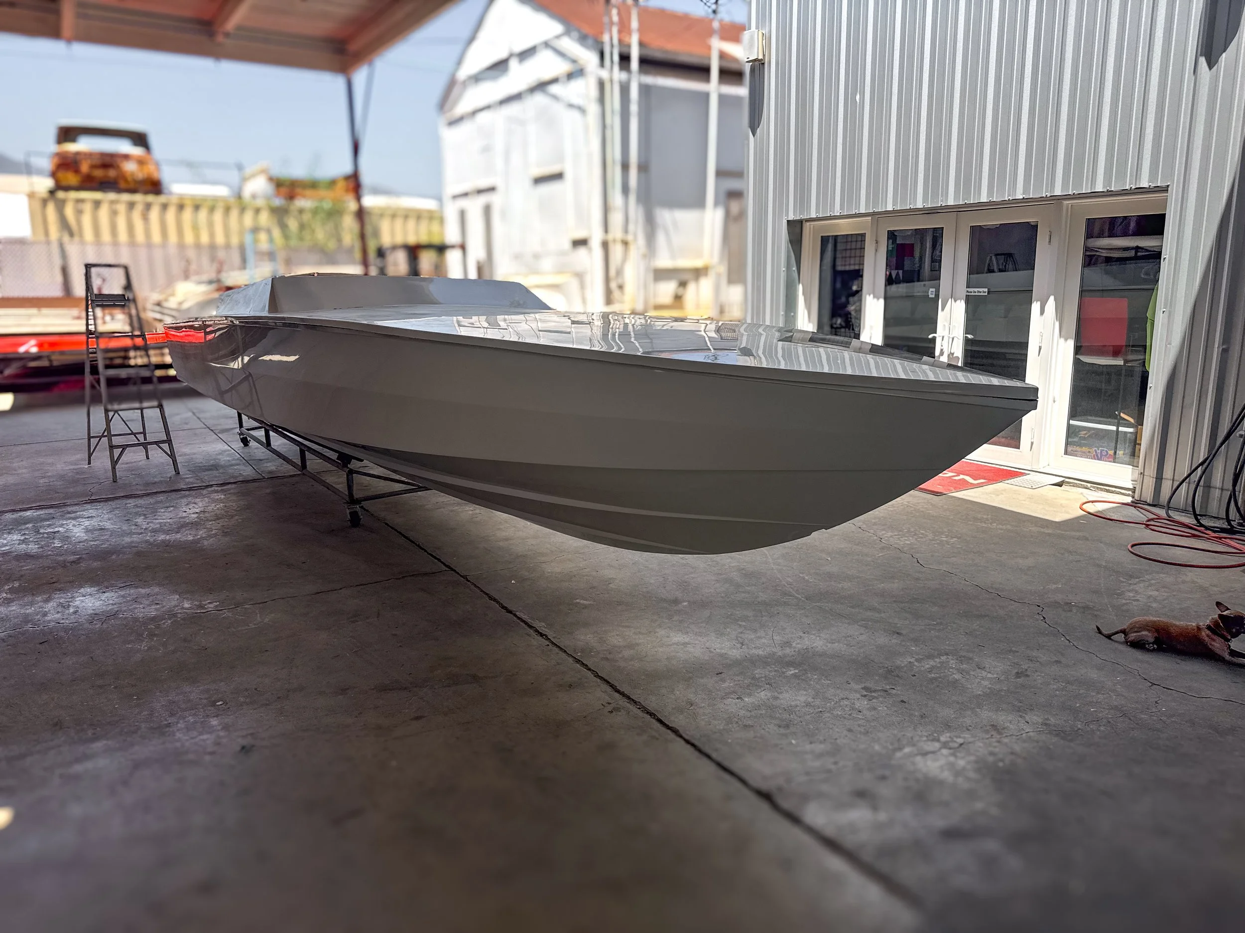 A boat on a trailer outside a warehouse or workshop with a gray metal exterior, some chairs, and a dog lying on the concrete floor.