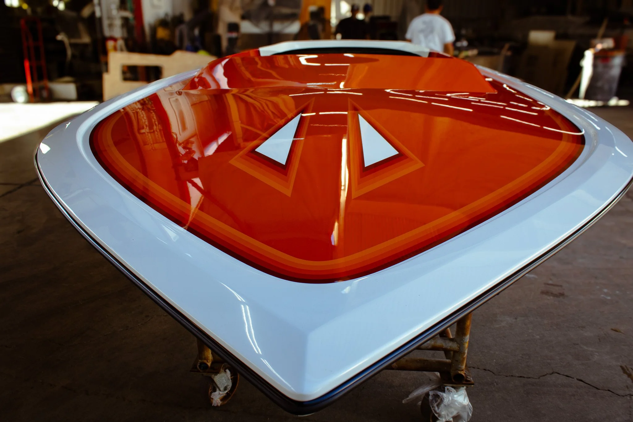 A close-up view of the hood of a white boat hull, featuring a large red and orange graphic with a stylized triangle design. The background shows a workshop or garage with people working.
