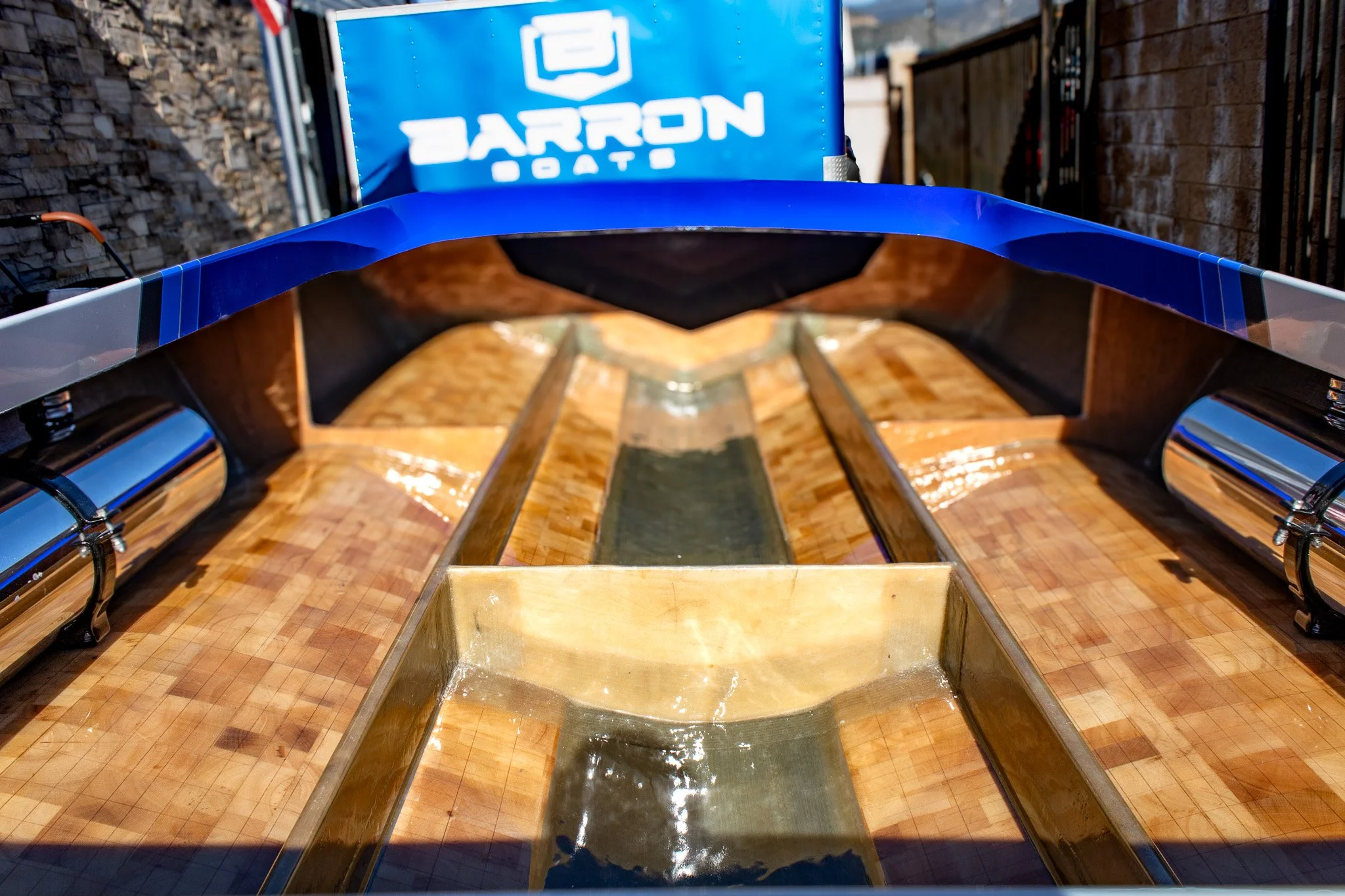 A boat hull with a blue and silver rail at the bow, positioned outdoors on a tiled surface, with a blue trailer in the background displaying a logo and the words 'BARRON BOATS'.