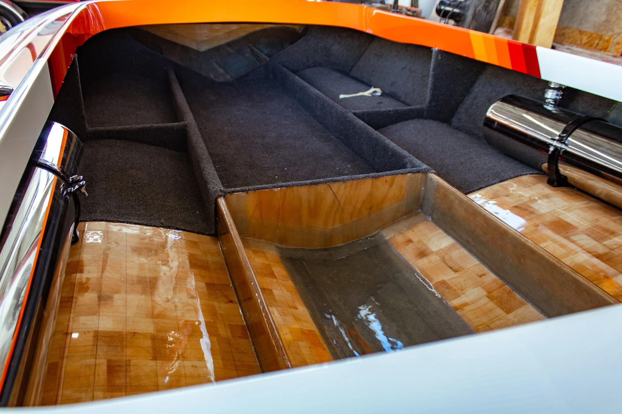 The interior of a boat hull showing an empty black carpeted cockpit with no seats, with a wooden floor underneath.