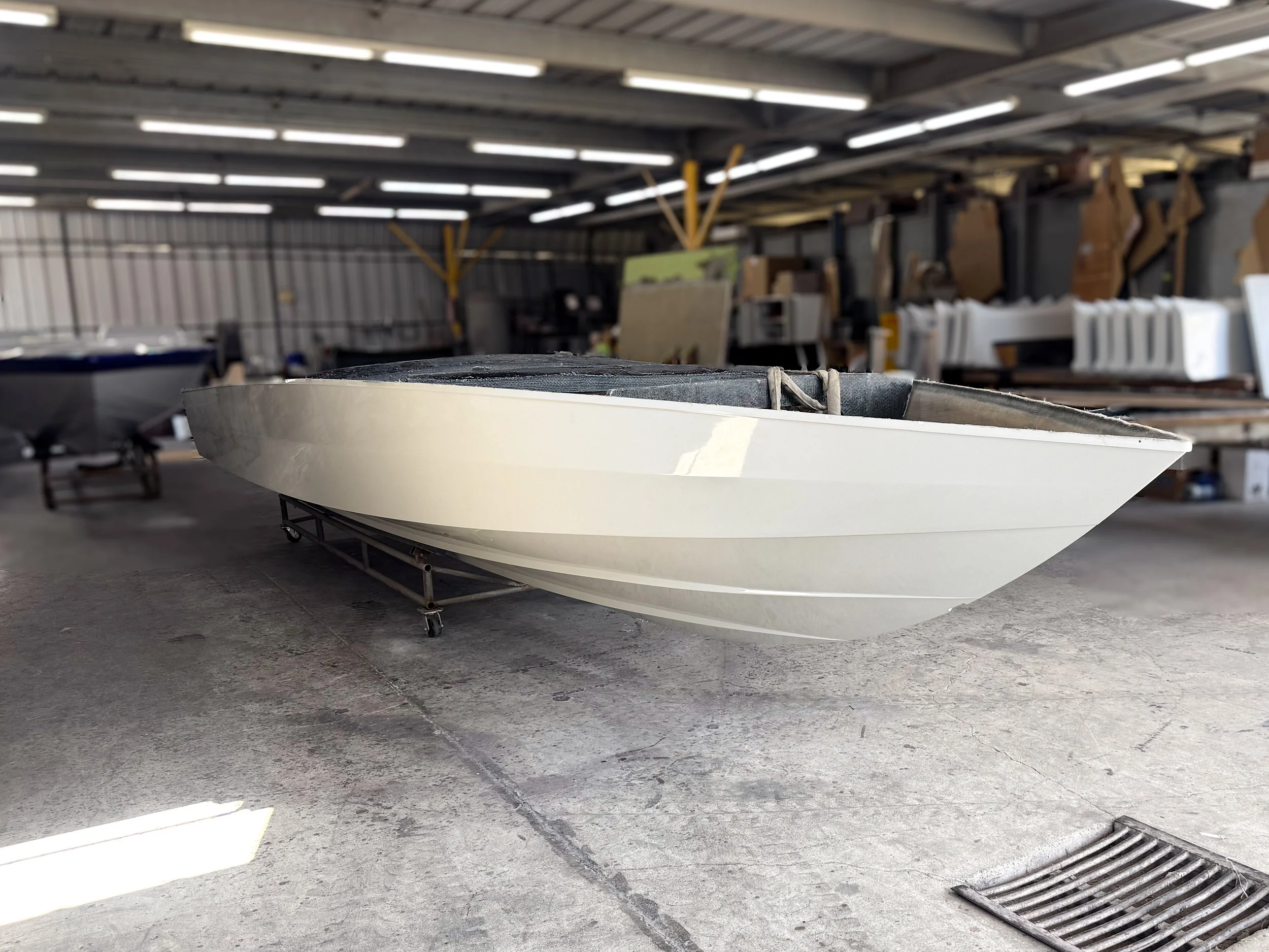 A white boat on a wheeled platform inside a warehouse or storage facility with metal beams, fluorescent lighting, and shelves with various materials.