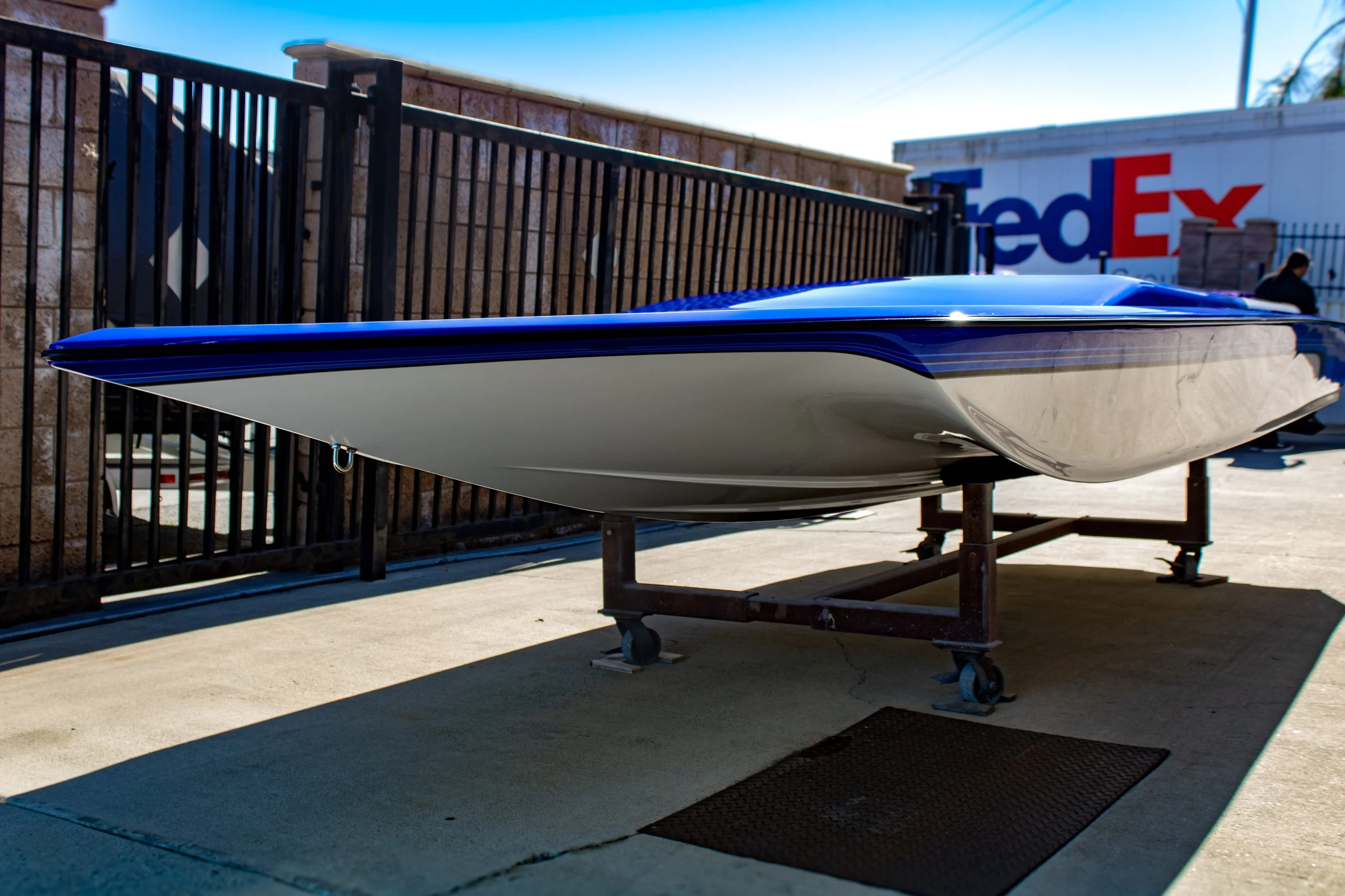 A small boat on a wheeled stand outdoors next to a black metal fence, with a FedEx truck in the background and a person walking nearby.
