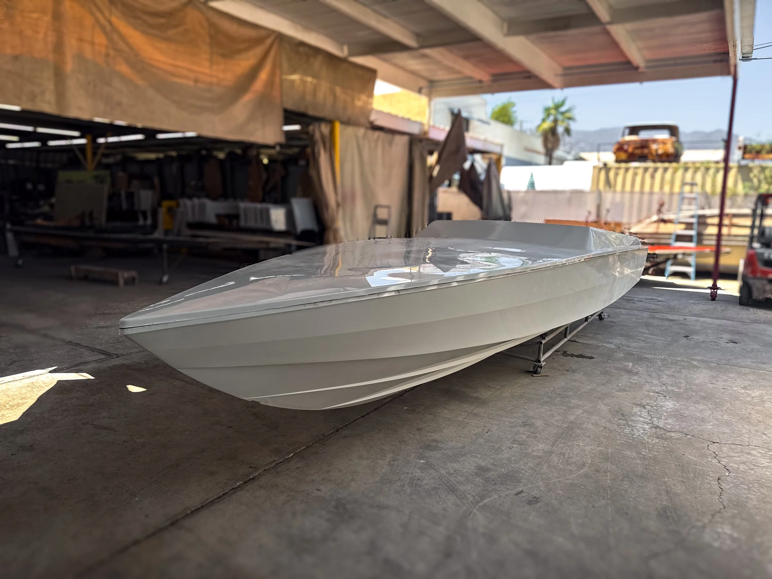 A white boat resting on a stand inside a garage or workshop with a concrete floor, wooden ceiling, and various equipment and materials in the background. Outdoors, a palm tree and mountains are visible in the distance.