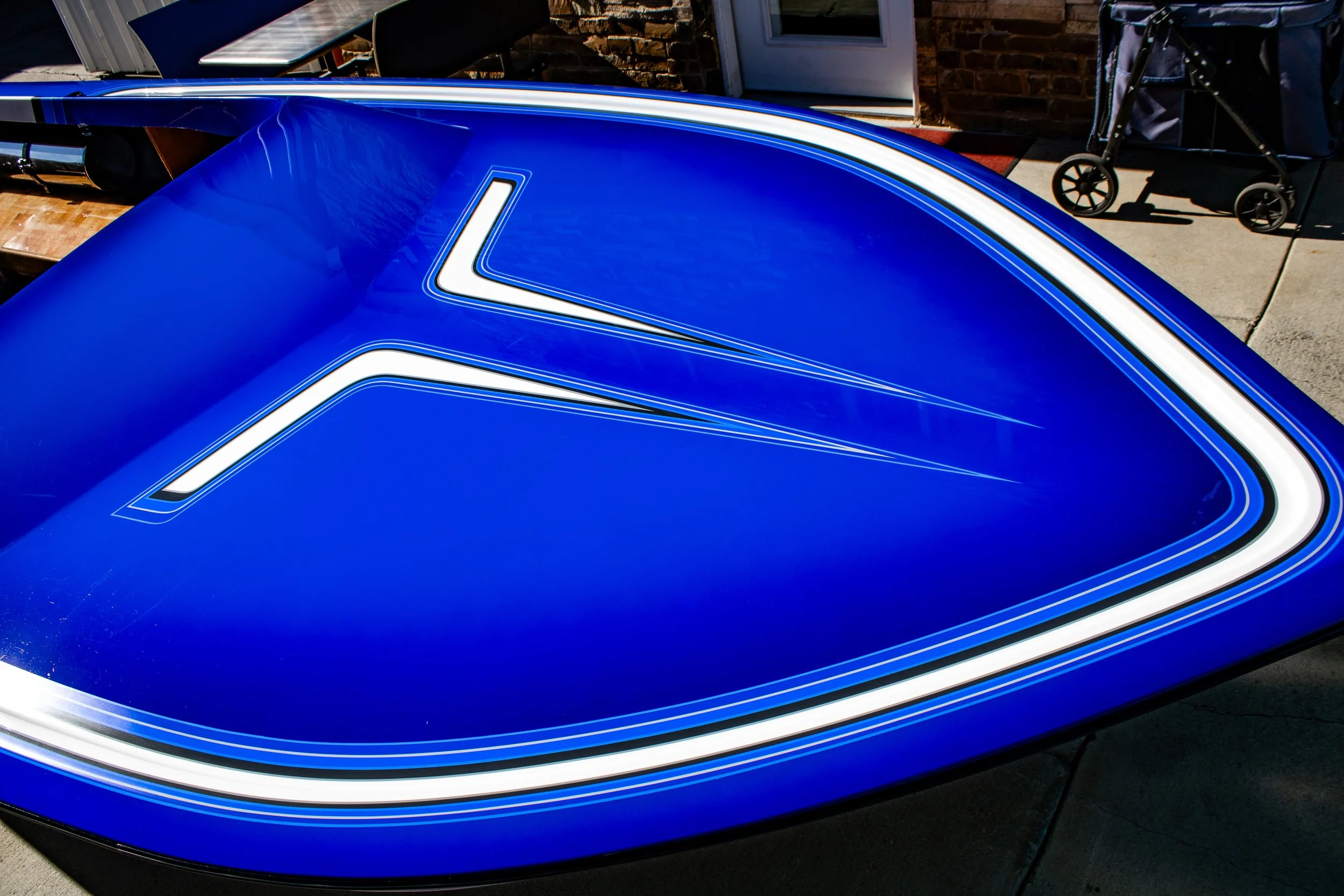 Close-up of the rear of a blue boat hull with white accent lines and a white outline on the trunk, parked outdoors near a brick building.