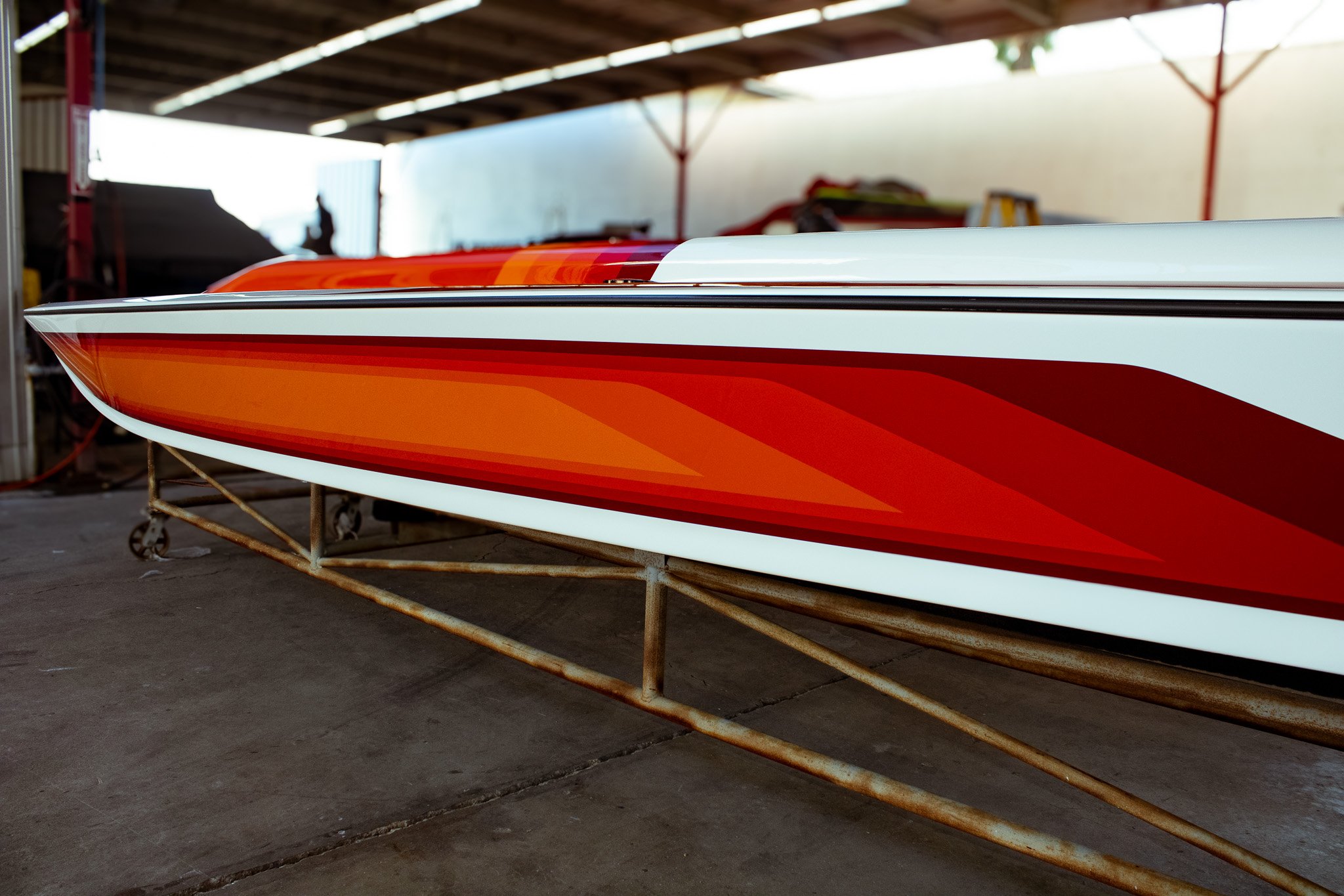 Close-up of a colorful boat with orange, red, and white design, stored on a metal rack in a workshop or storage area with a partially covered roof.