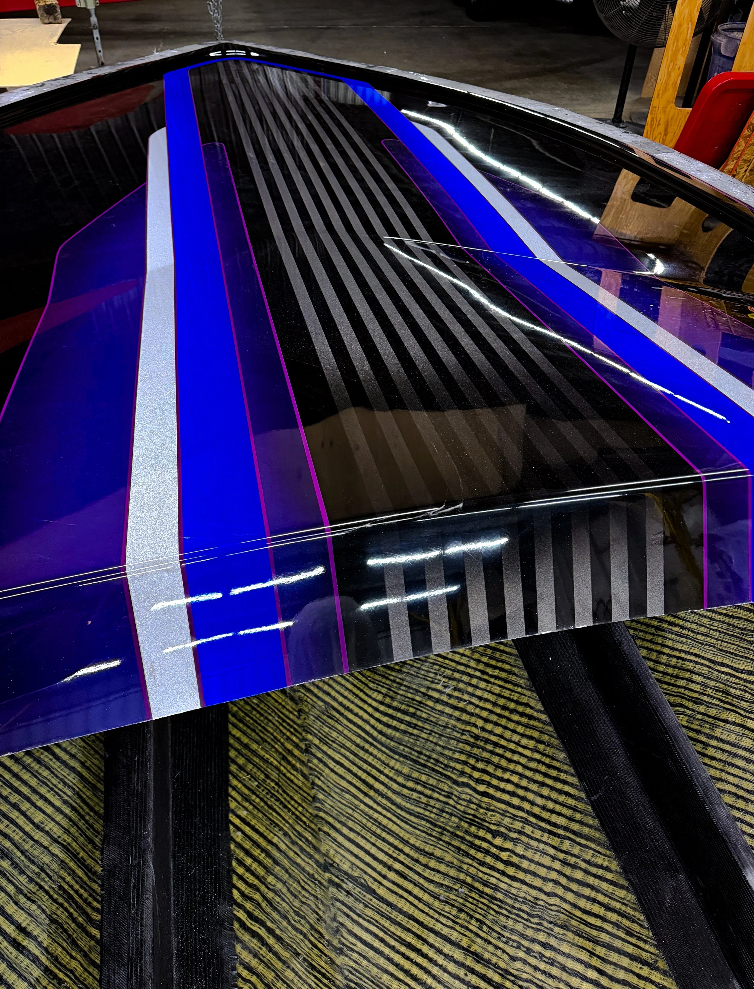 A boat hull with a custom paint job featuring black, white, blue, and purple stripes and neon accents, reflecting overhead lighting.