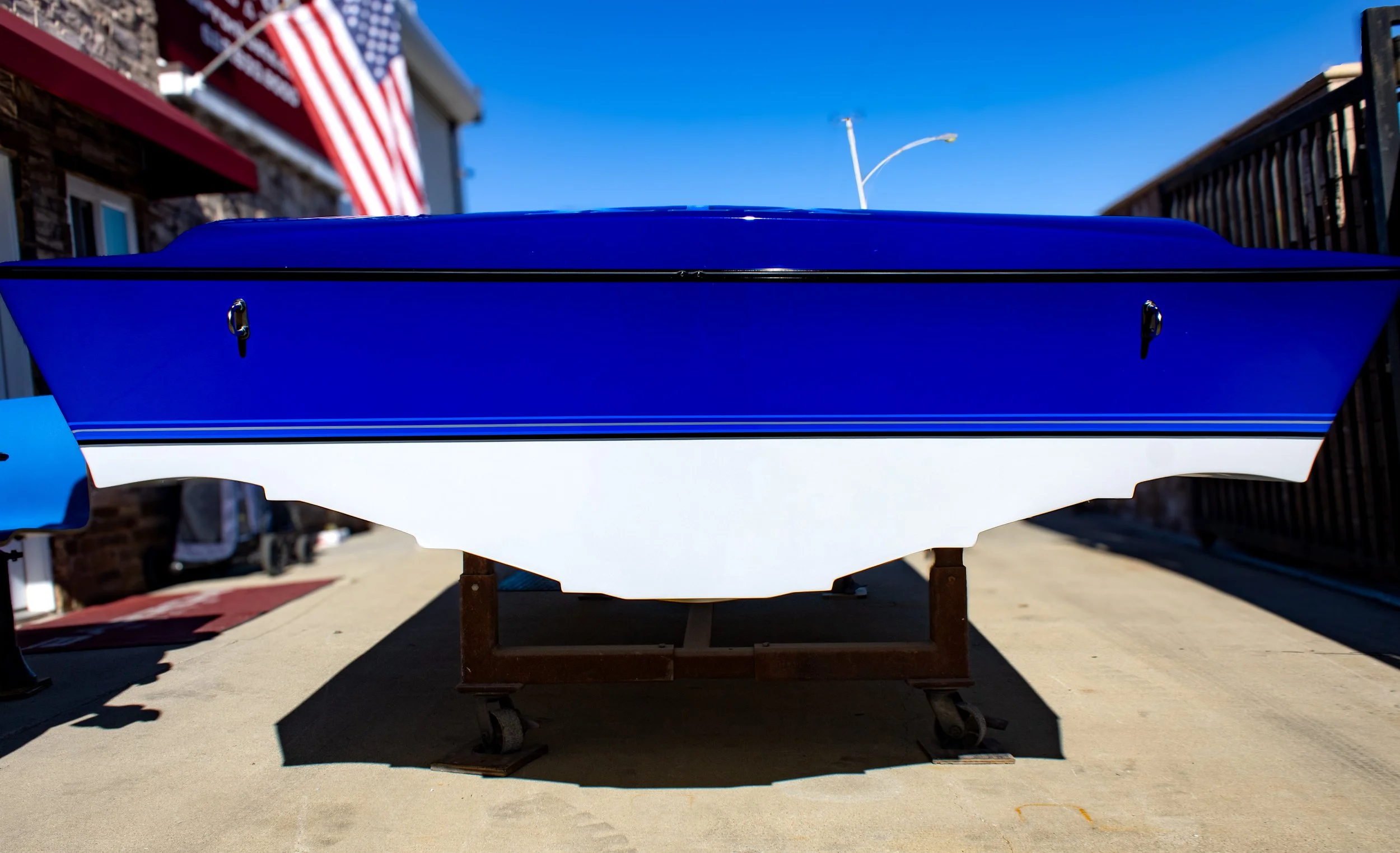 A boat with a blue top and white bottom on a wooden stand outdoors, with a parking lot, buildings, and American flags in the background under a clear blue sky.