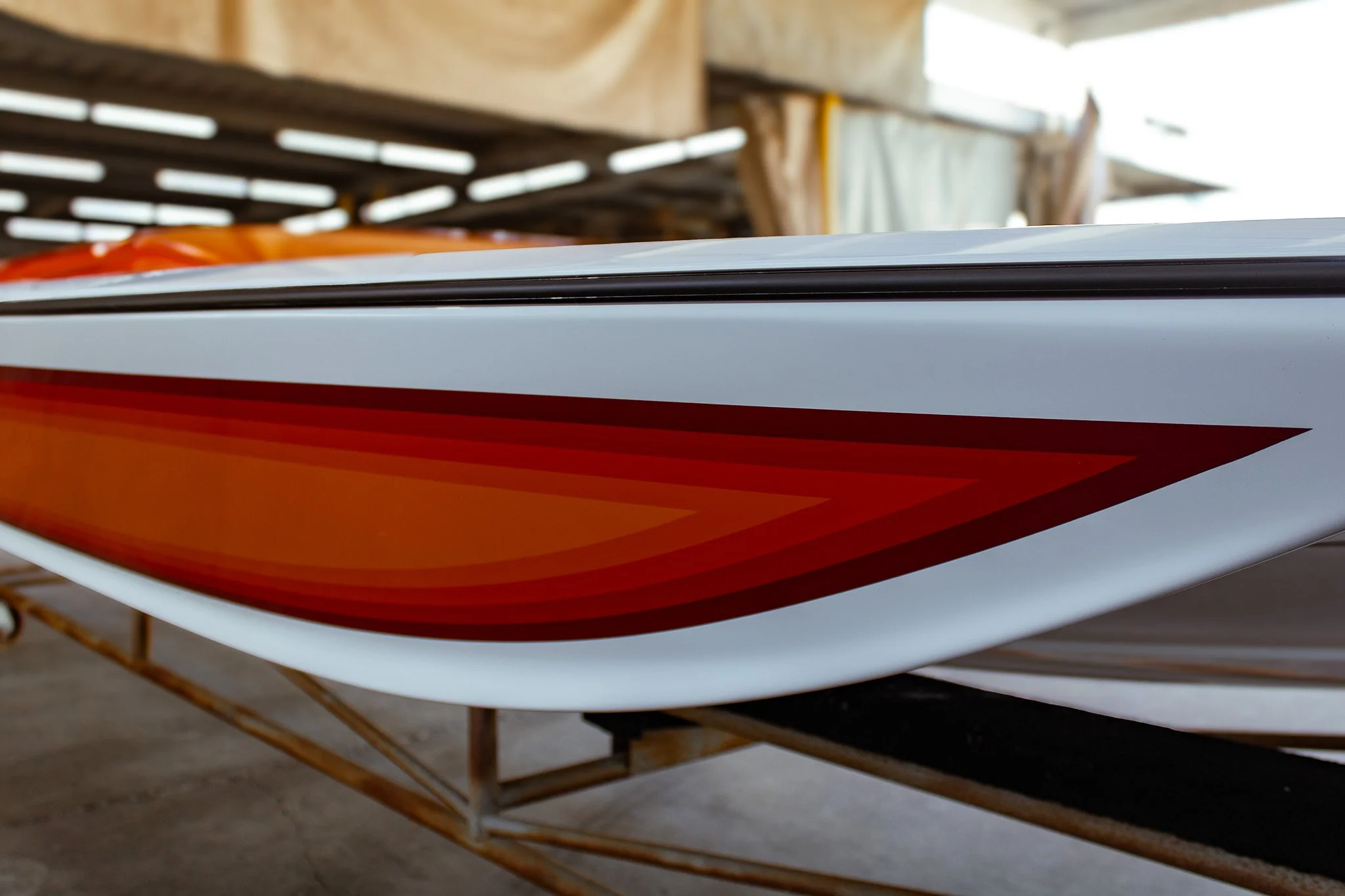 A close-up side view of a colorful boat hull, showing orange, red, and white sections, resting on a pallet in an indoor storage area with wooden beams.