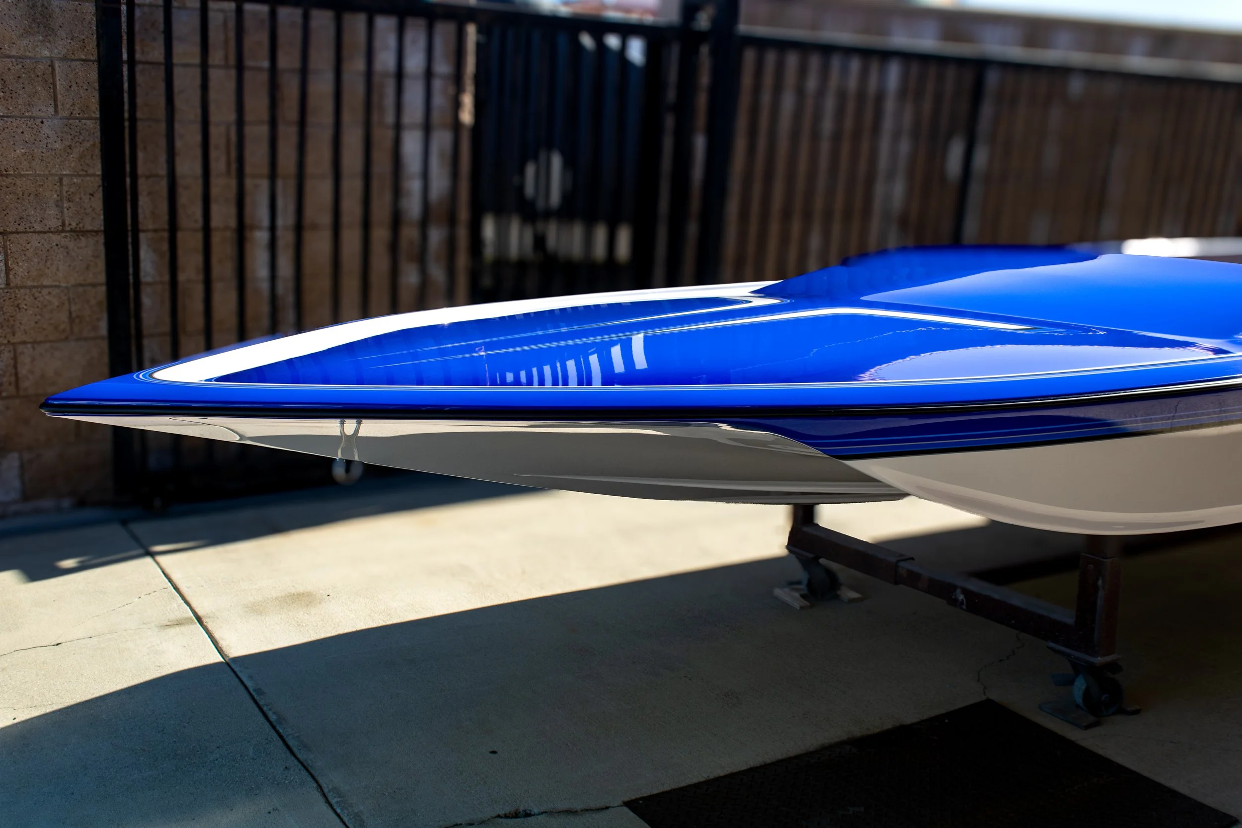 A blue and white boat stored on a wheeled boat trailer on a concrete patio, with a brick wall and black metal fence in the background.