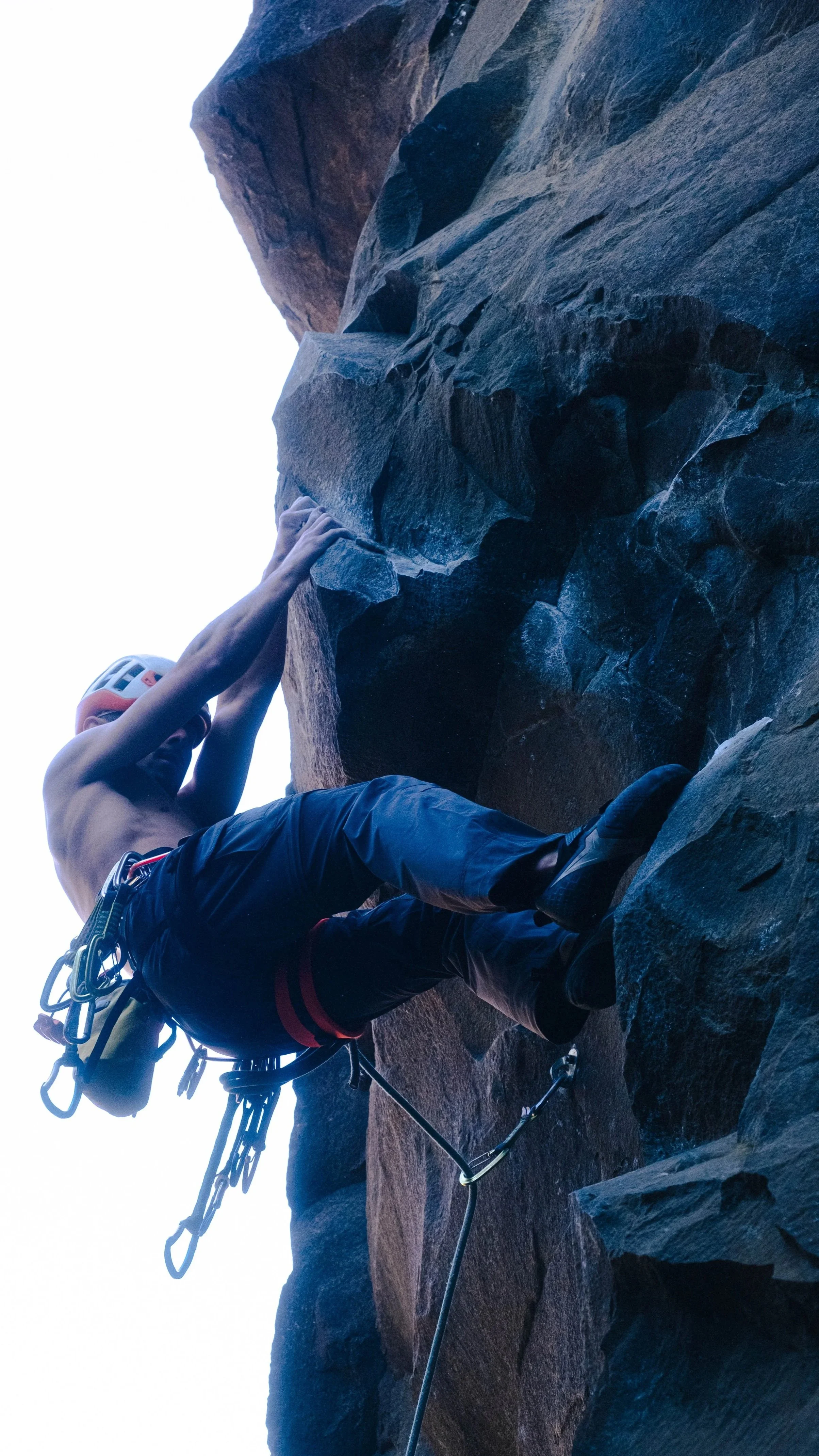 Escalador sin camisa en una roca, usando arnés y casco, escalando una pared de roca en un ambiente al aire libre.