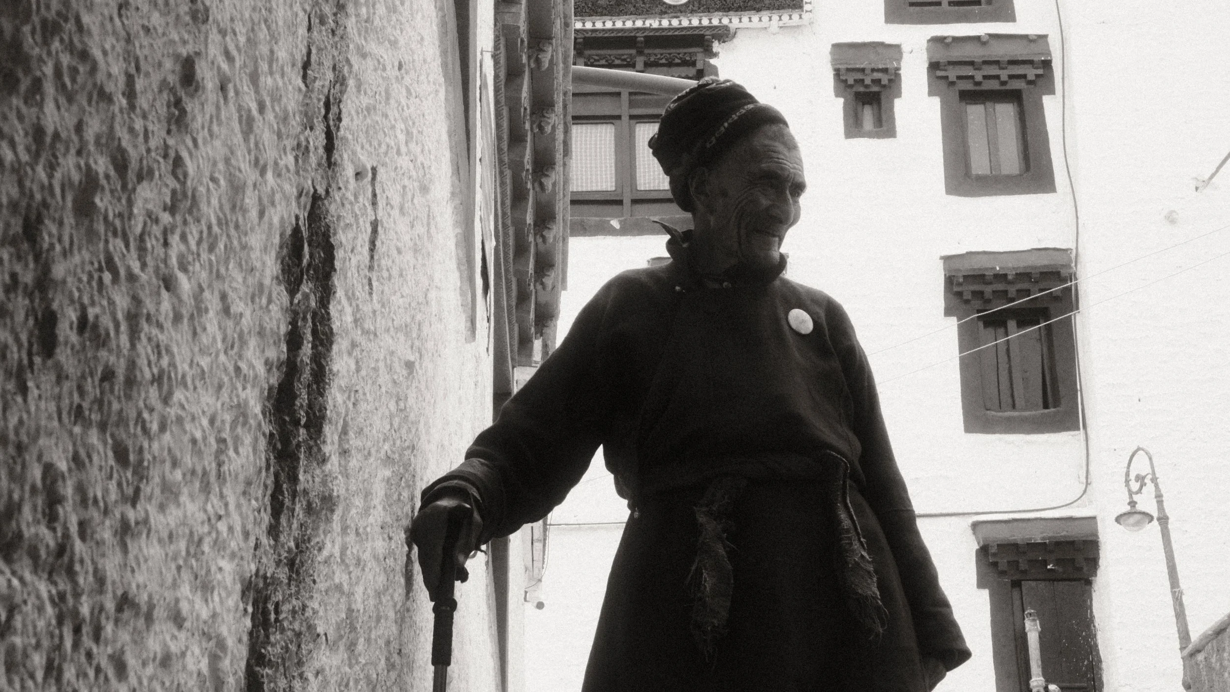 Anciana con bastón caminando por una calle estrecha de piedra, con edificios tradicionales en el fondo.