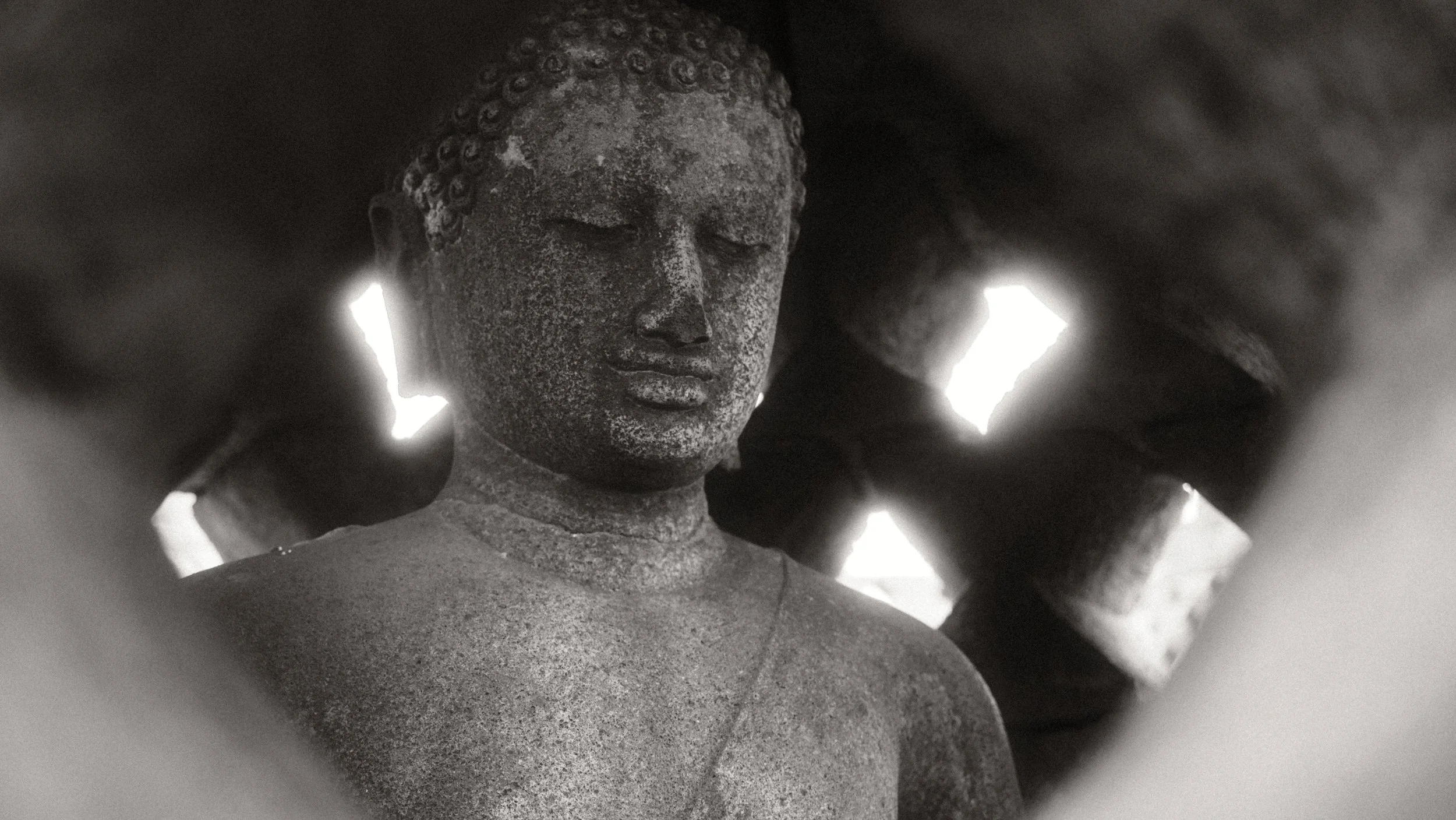 Imagen en blanco y negro de una estatua de Buda con ojos cerrados, en un espacio con paredes perforadas que dejan pasar luz natural.