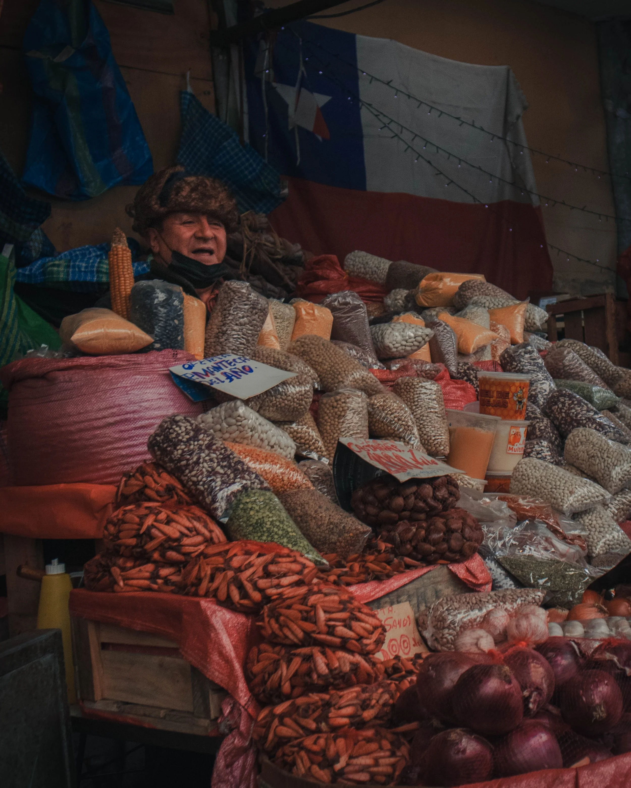 Vendedor en puesto de mercado vendiendo legumbres, arroz y cebolla en bolsas y sacos.