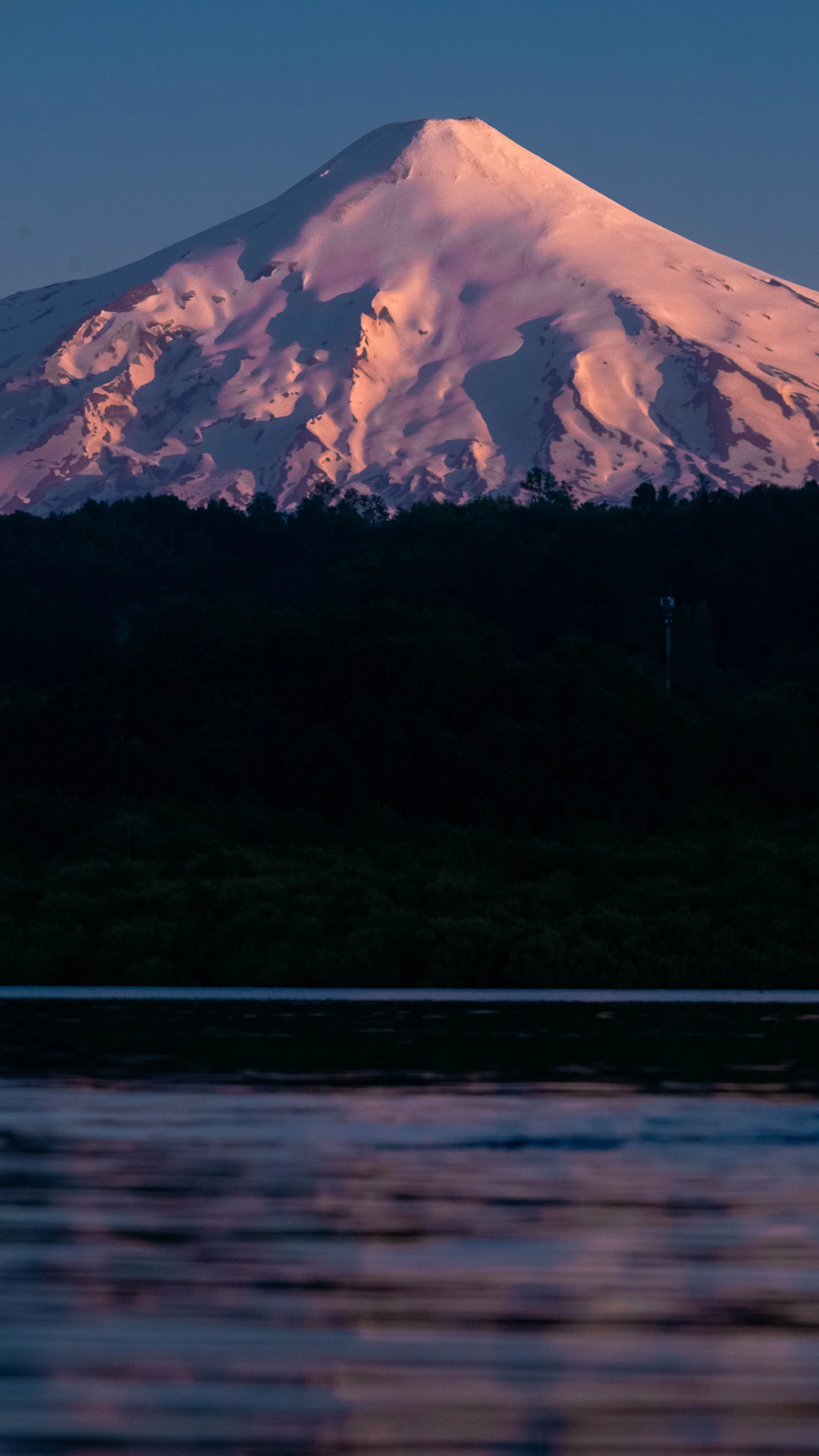 Monte con cumbres nevadas y cielo claro, reflejado en un cuerpo de agua en primer plano.