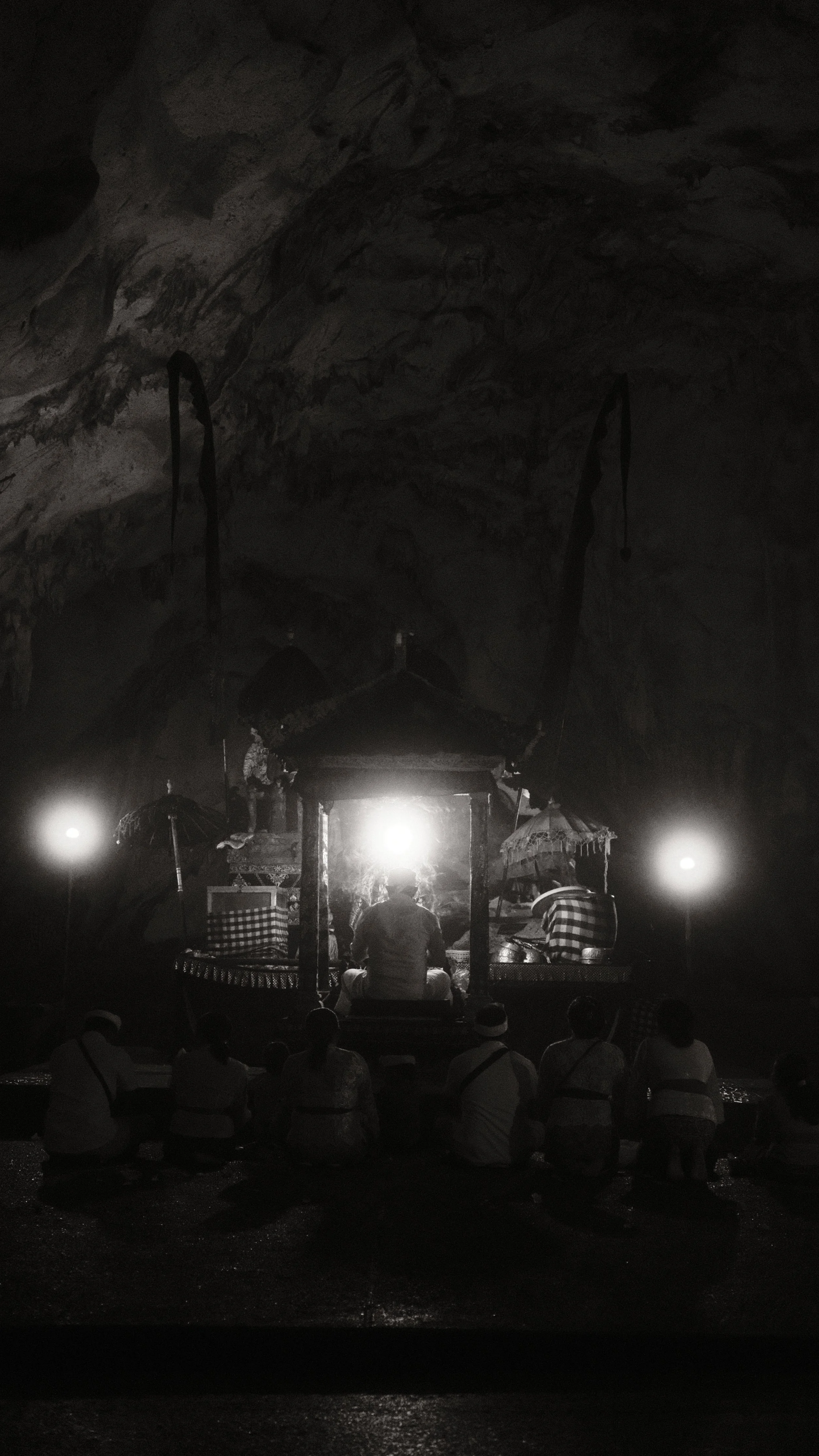 Escena en una cueva con personas participando en una ceremonia o ritual, con iluminación tenue y un altar en el centro con un hombre sentado frente a él, rodeado de paraguas y objetos tradicionales.