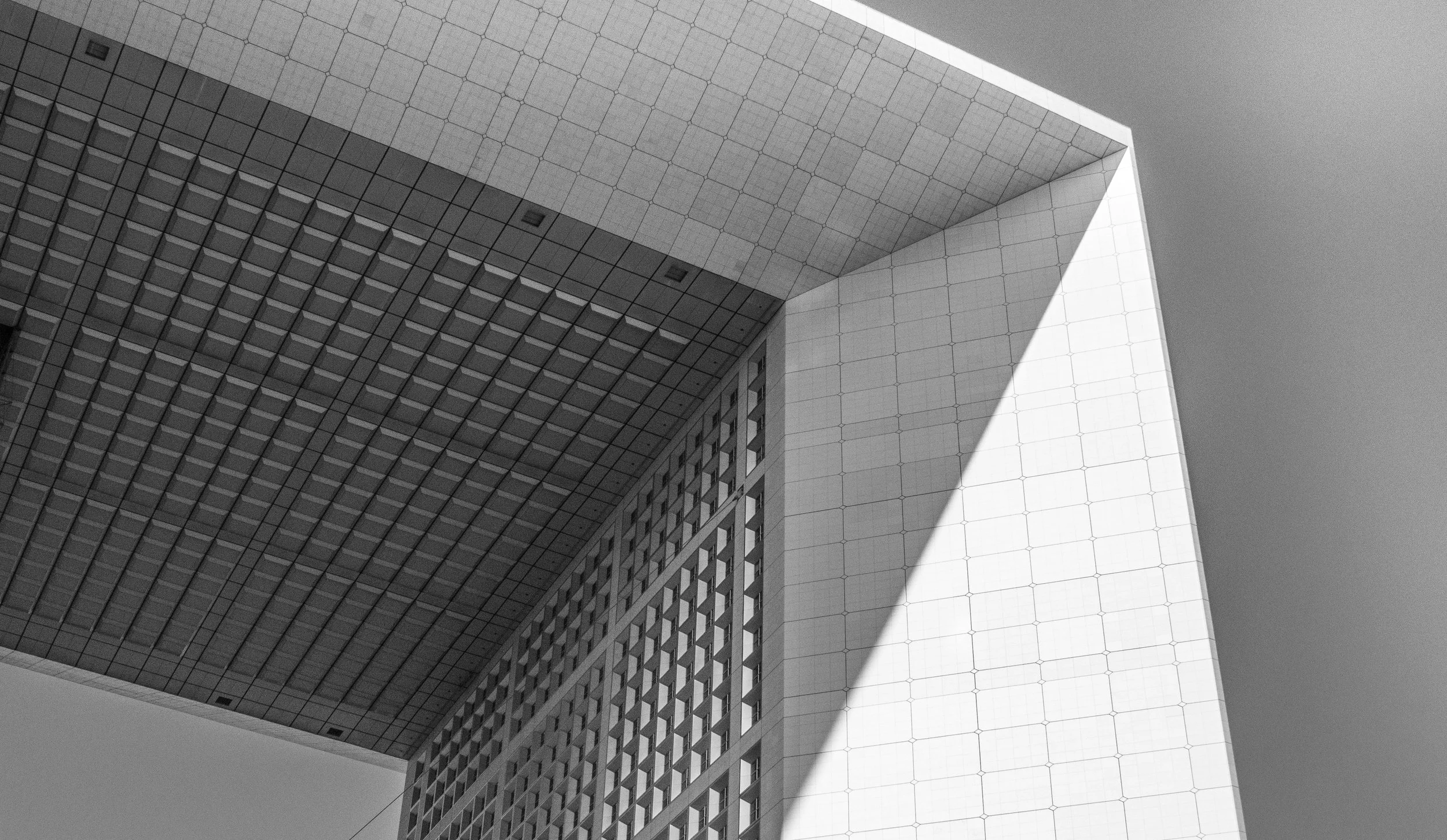 Black and white photo of a modern building with geometric patterns and shadow play, showing a large overhanging structure and grid-like facade.