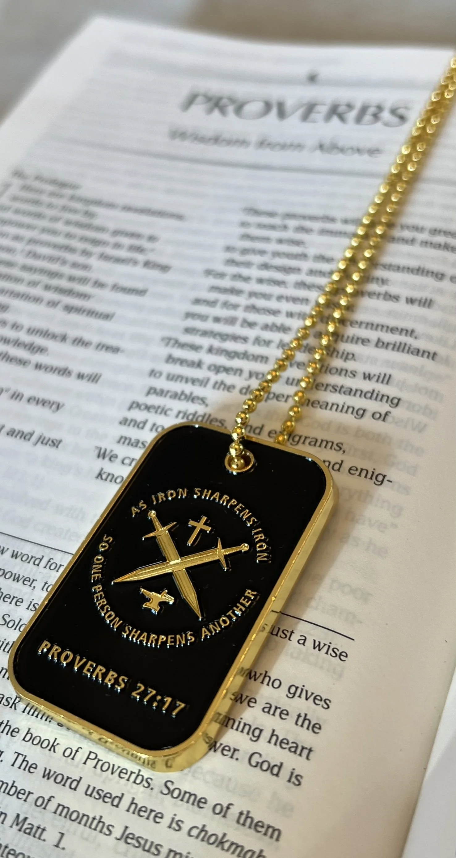 A black and gold necklace with a pendant featuring crossed swords, a small cross, and a scroll with the quote from Proverbs 27:17, placed on a Bible page.