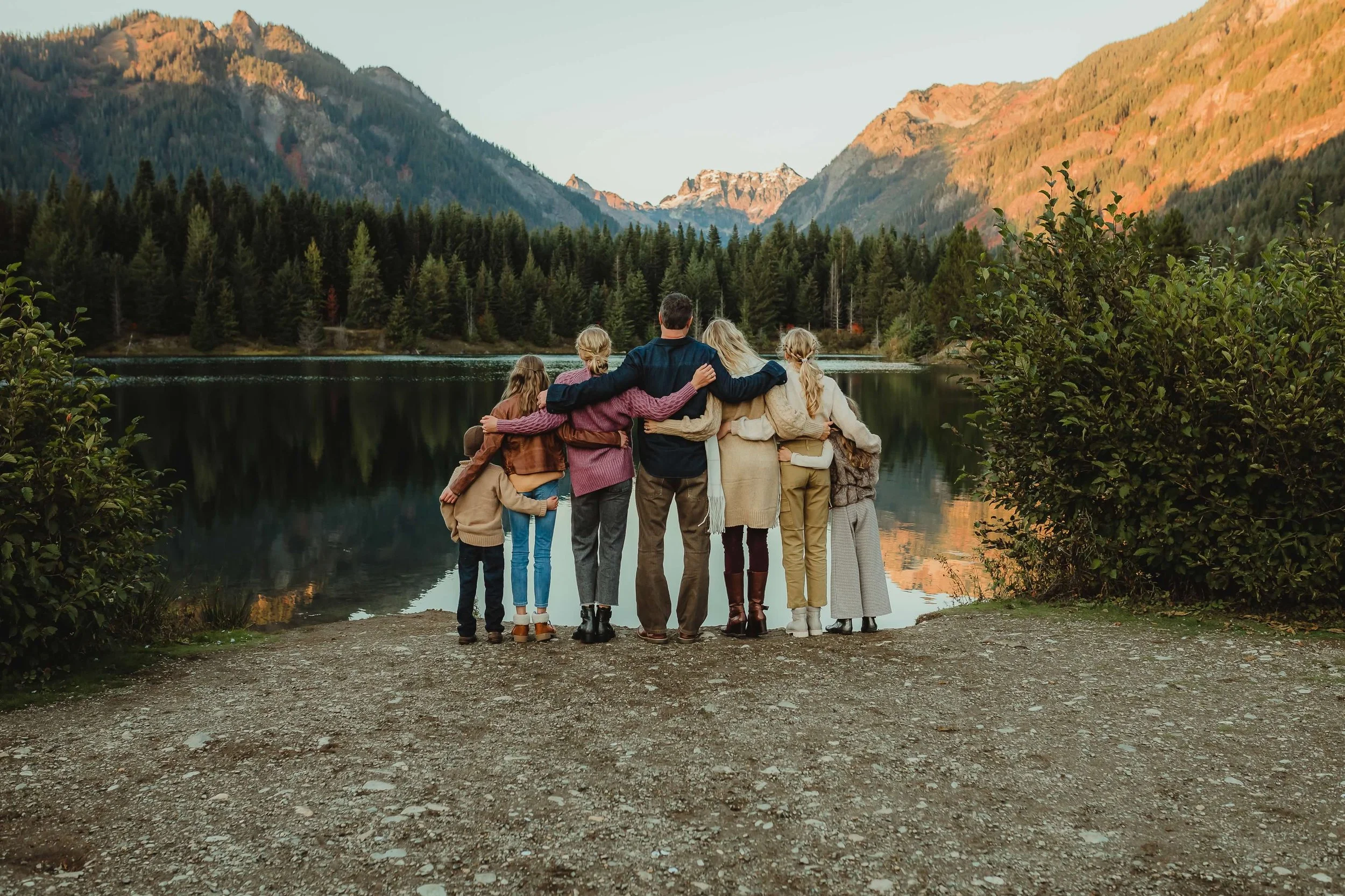 Family of seven standing by a lake at sunset, embracing each other, with mountains and forest in the background.