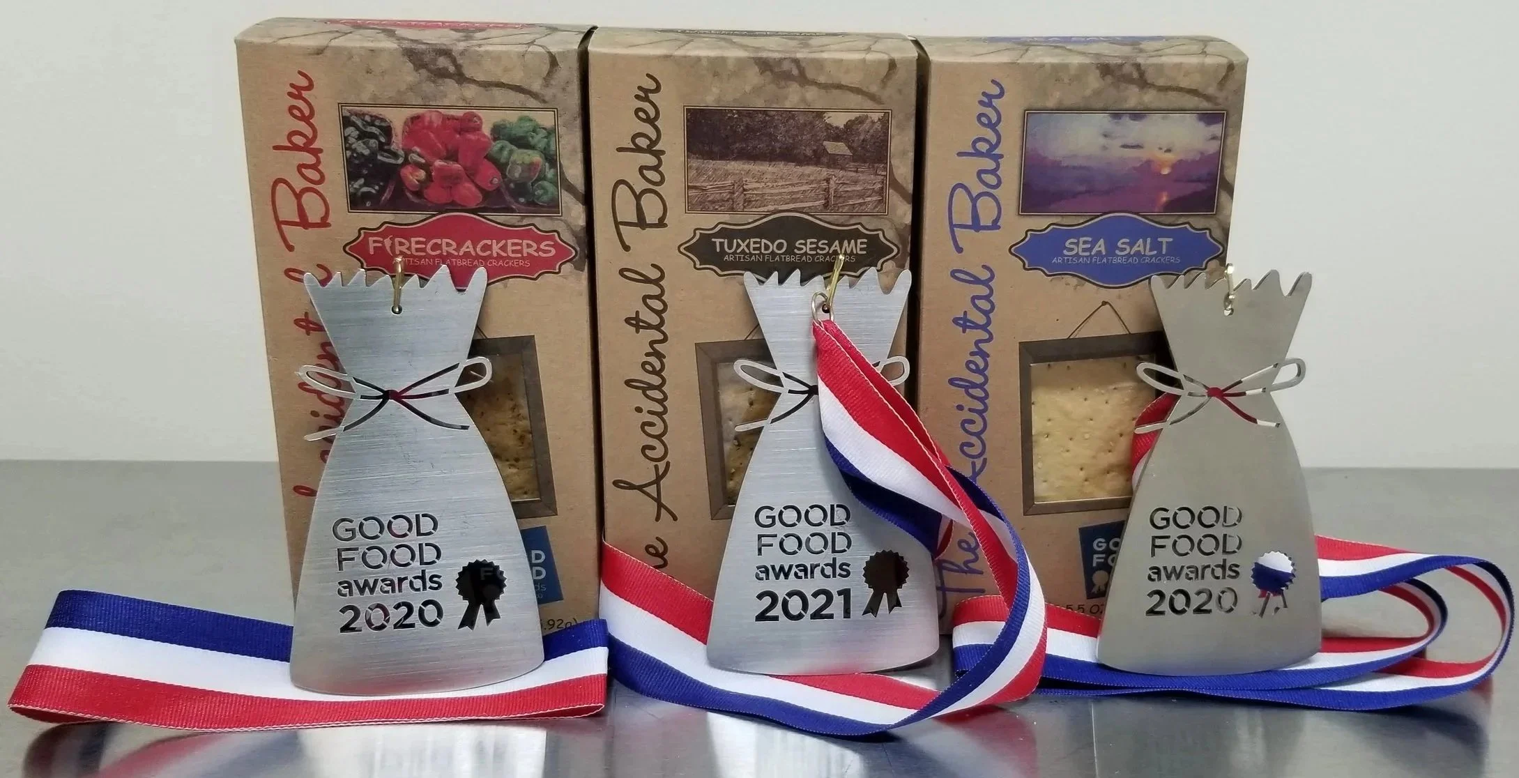 Three boxes of artisan flatbread crackers: Firecrackers, Tuxedo Sesame, and Sea Salt. Leaned against each box is a medal with a ribbon reading 'Good Food Awards 2020' or '2021'.