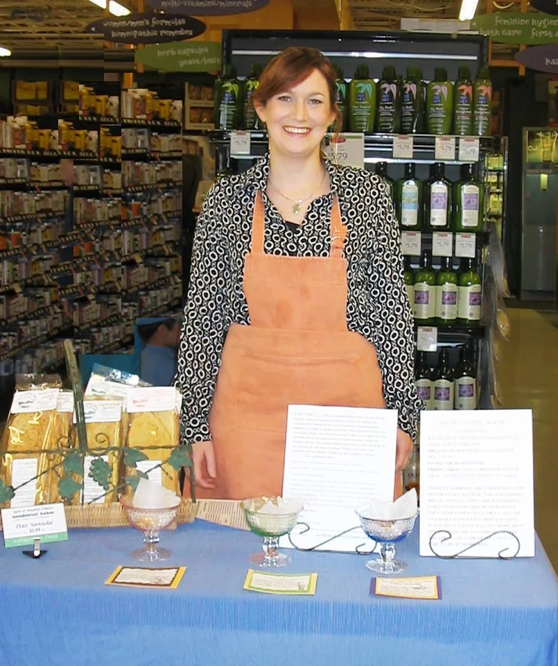 A woman in a patterned blouse and peach apron standing behind a table with bowls and informational signs in a store aisle filled with various products.