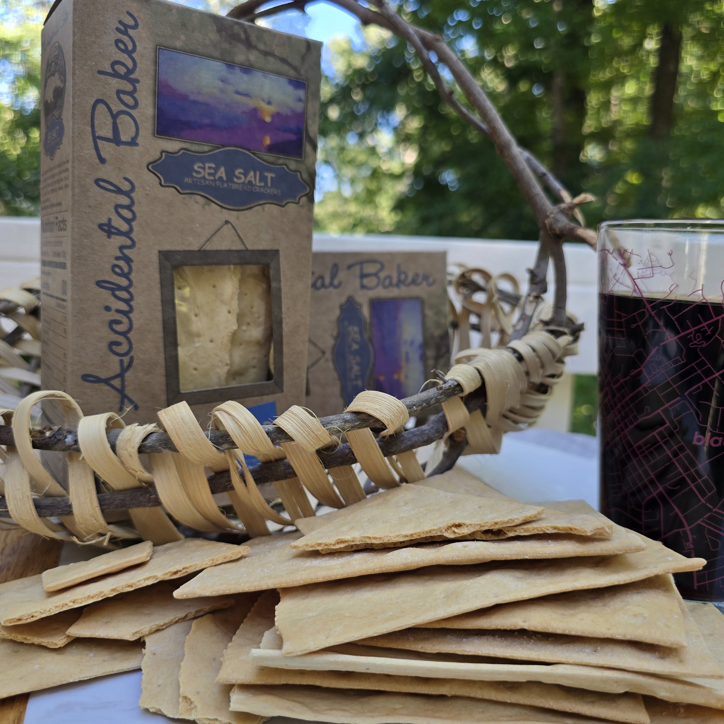 A stack of Sea Salt flatbread crackers showing broken crackers and the layers within each crispy cracker in front of a box of sea salt artisanal flatbread crackers and a glass of dark beer on a patio table.