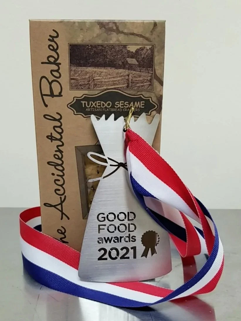 A silver award trophy with a red, white, and blue ribbon draped over it, placed in front of a box labeled 'Tuxedo Sesame' and 'The Accredited Baker,' with a small illustrations of a rustic barn and a landscape in the background.