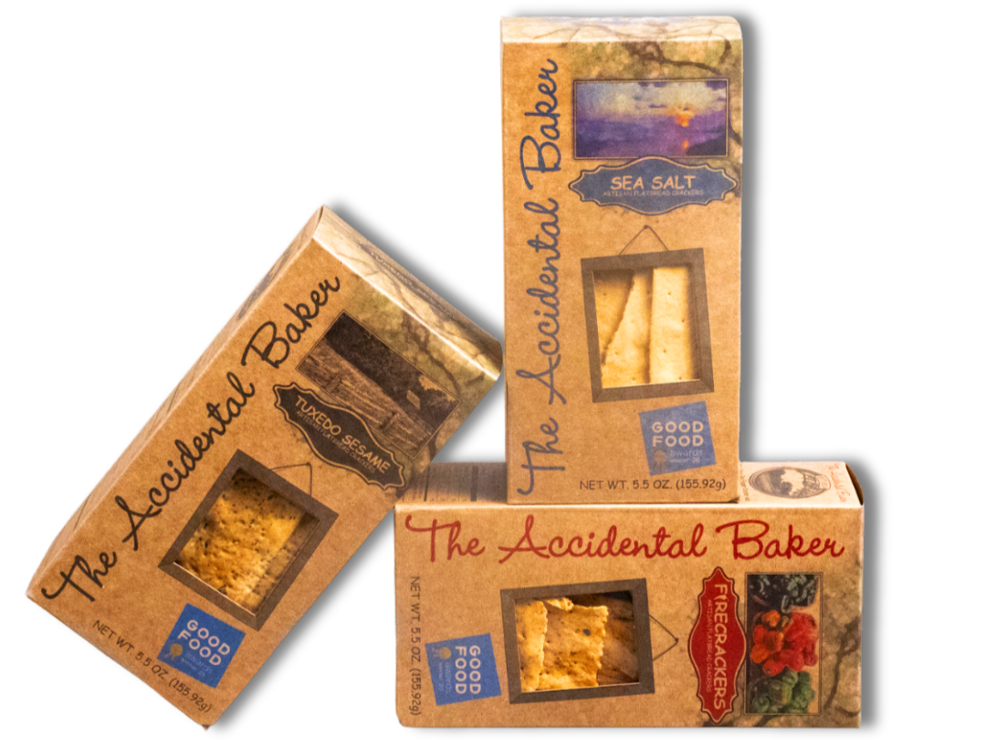 Three boxes of The Accidental Baker sea salt snack crackers, one box upright and one box tilted, all featuring transparent windows showing the crackers inside, with a black background.