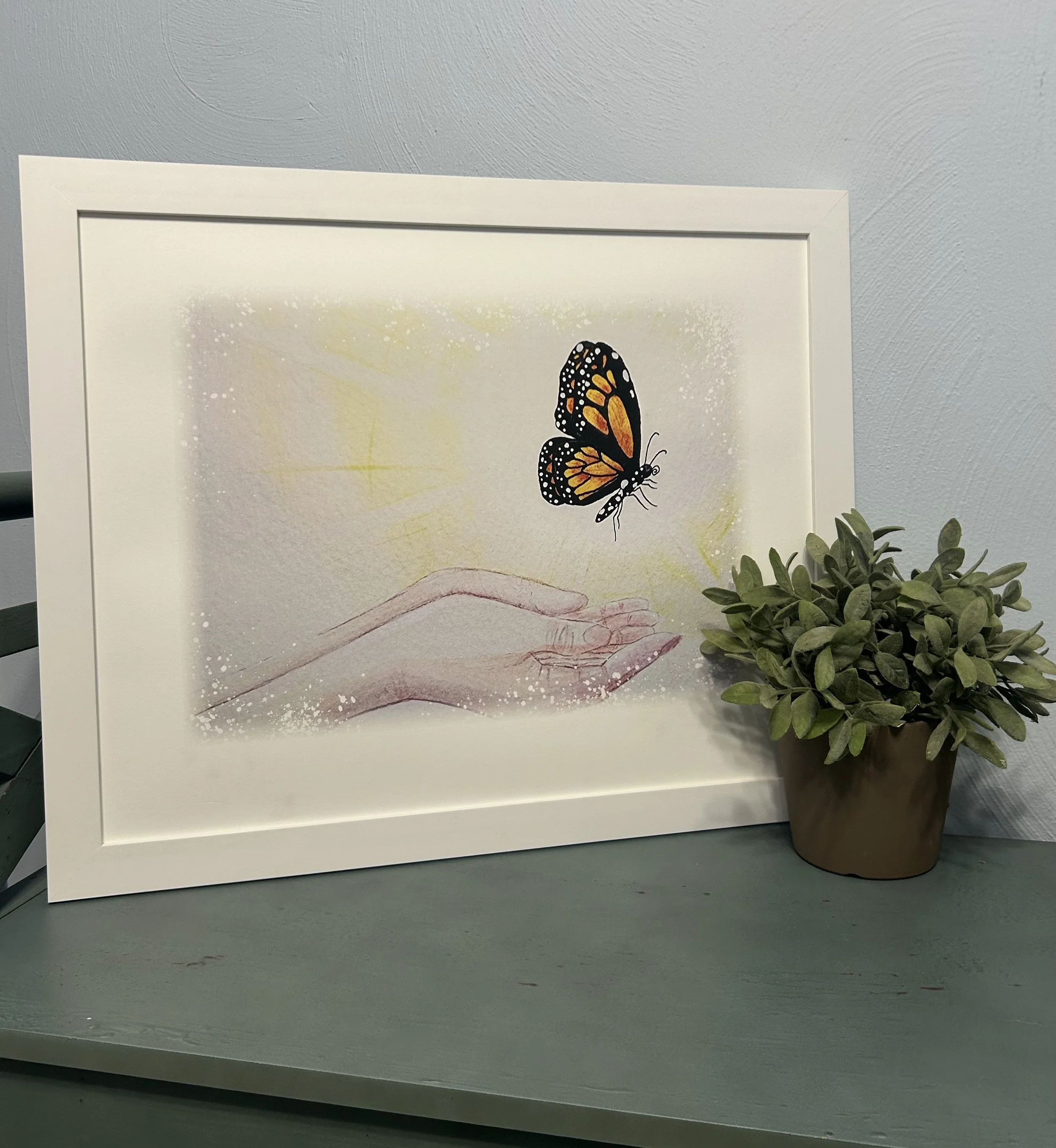 Framed artwork showing a hand reaching out with a butterfly flying above it, placed next to a potted green plant, on a green surface against a white wall.