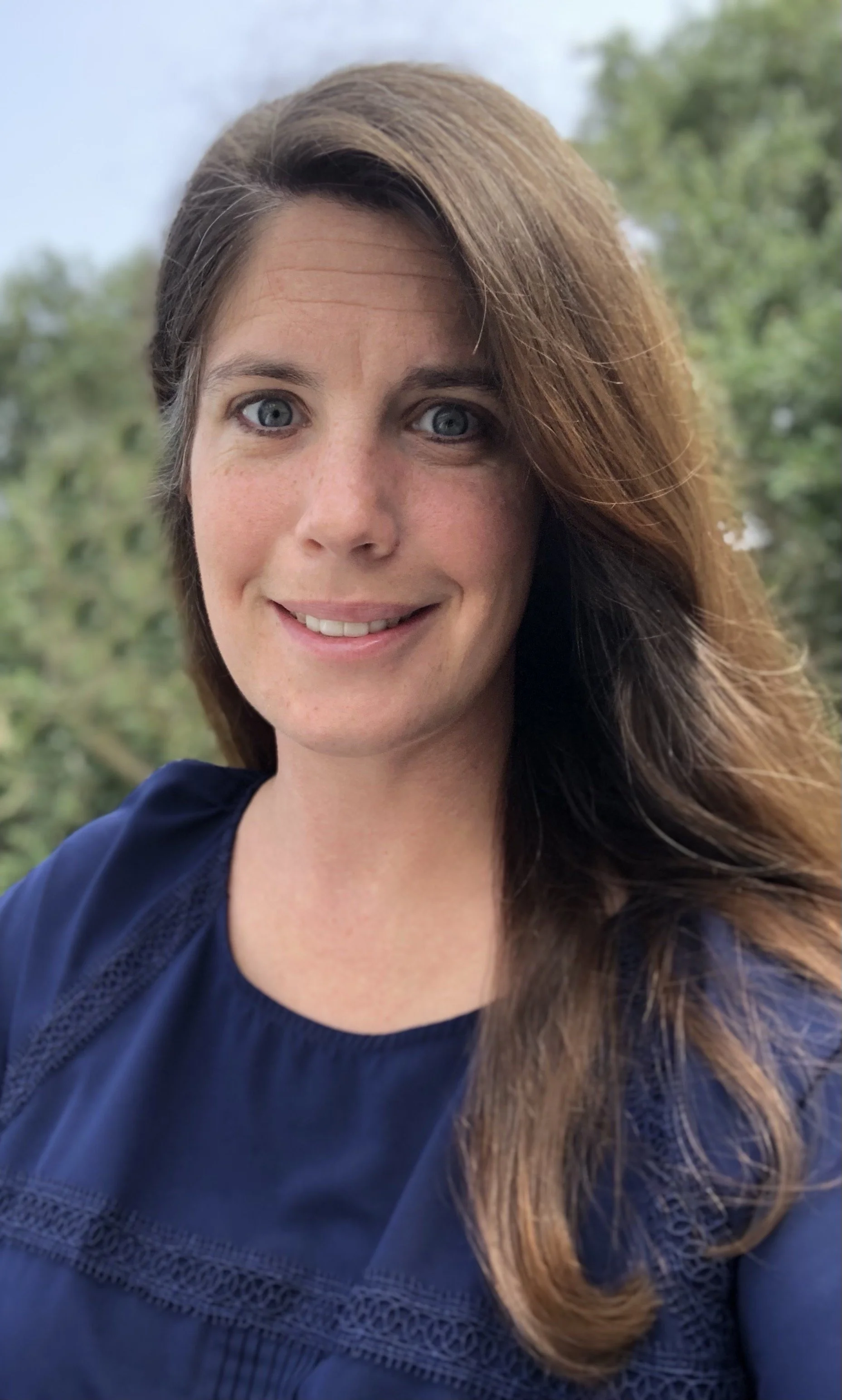 A woman with long brown hair and blue eyes smiling outdoors with green trees in the background.