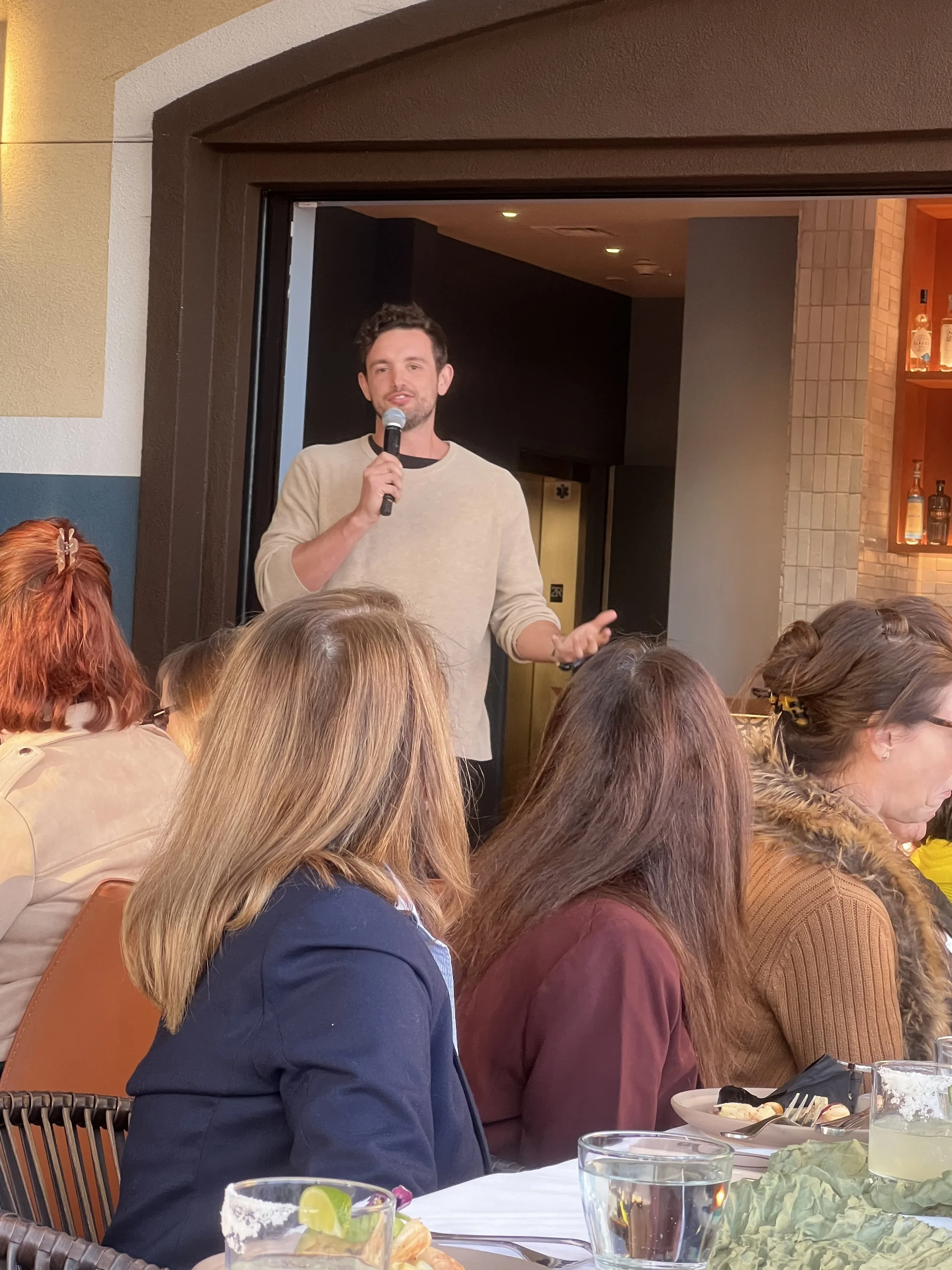 A man standing with a microphone talking to an audience at a restaurant or cafe, with several women sitting at a table.