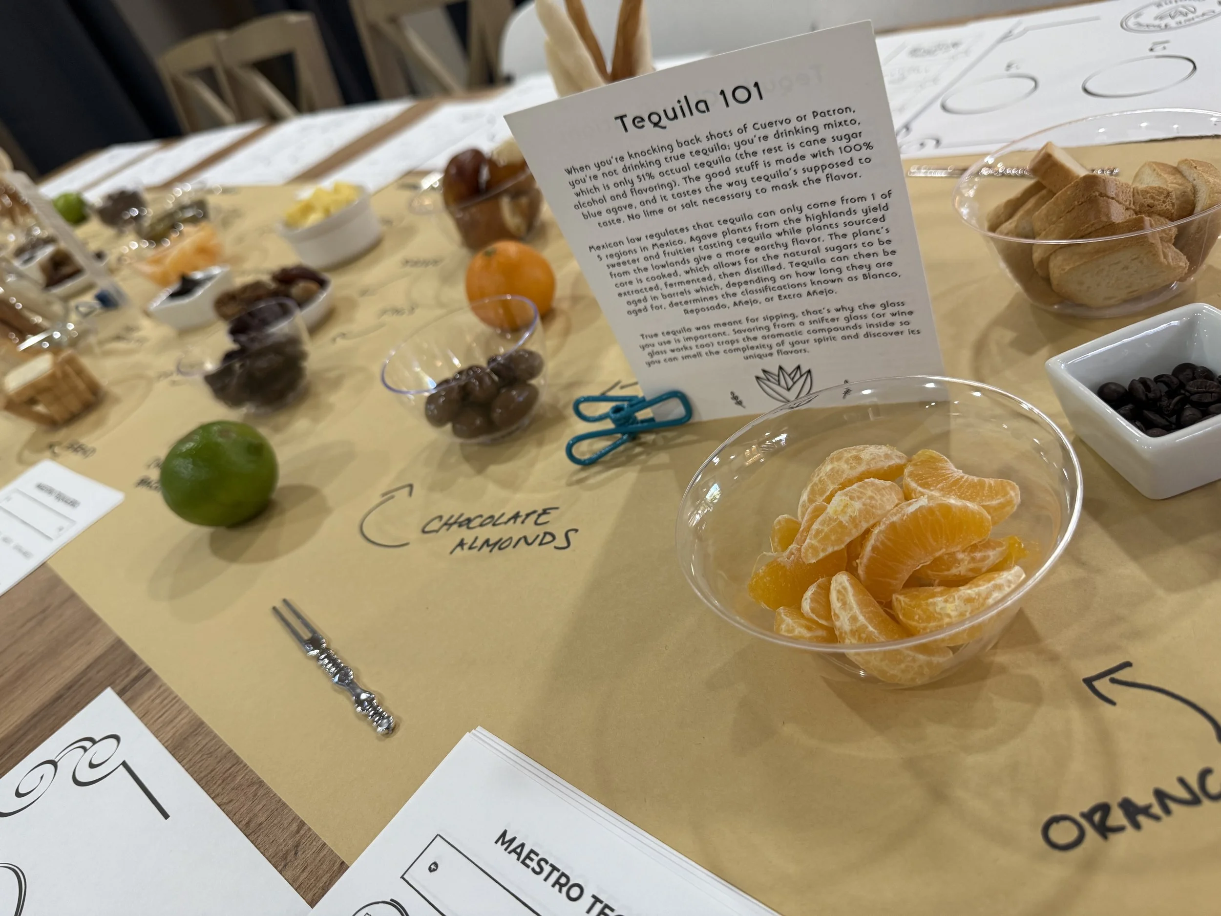 Table display with various ingredients and a sign titled "Tequila 101" explaining tequila types, along with bowls containing oranges, chocolate almonds, and other snacks, and handwritten signs indicating different flavors.