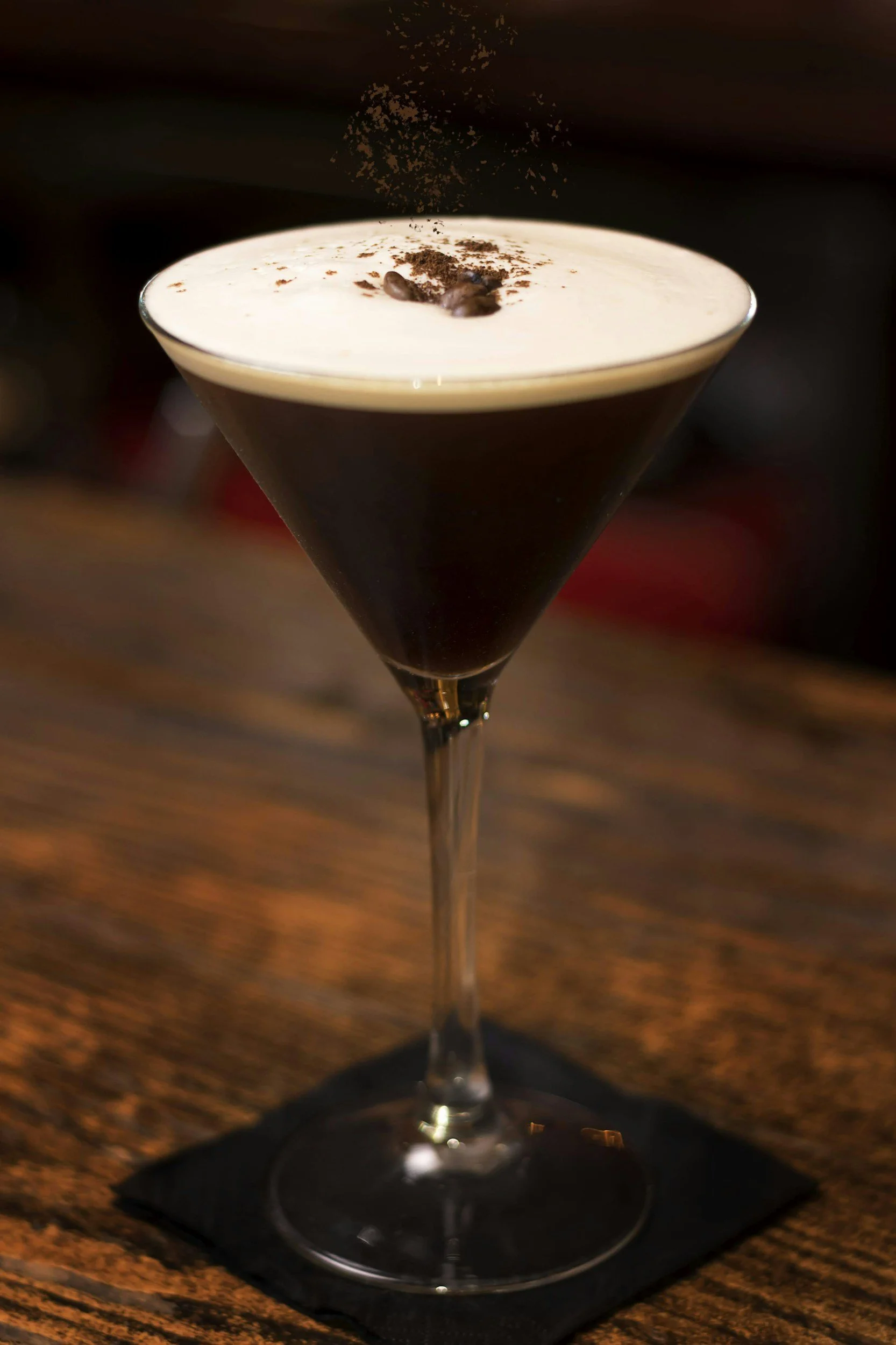 A dark coffee cocktail in a martini glass topped with whipped cream and cocoa powder, sitting on a wooden bar counter.