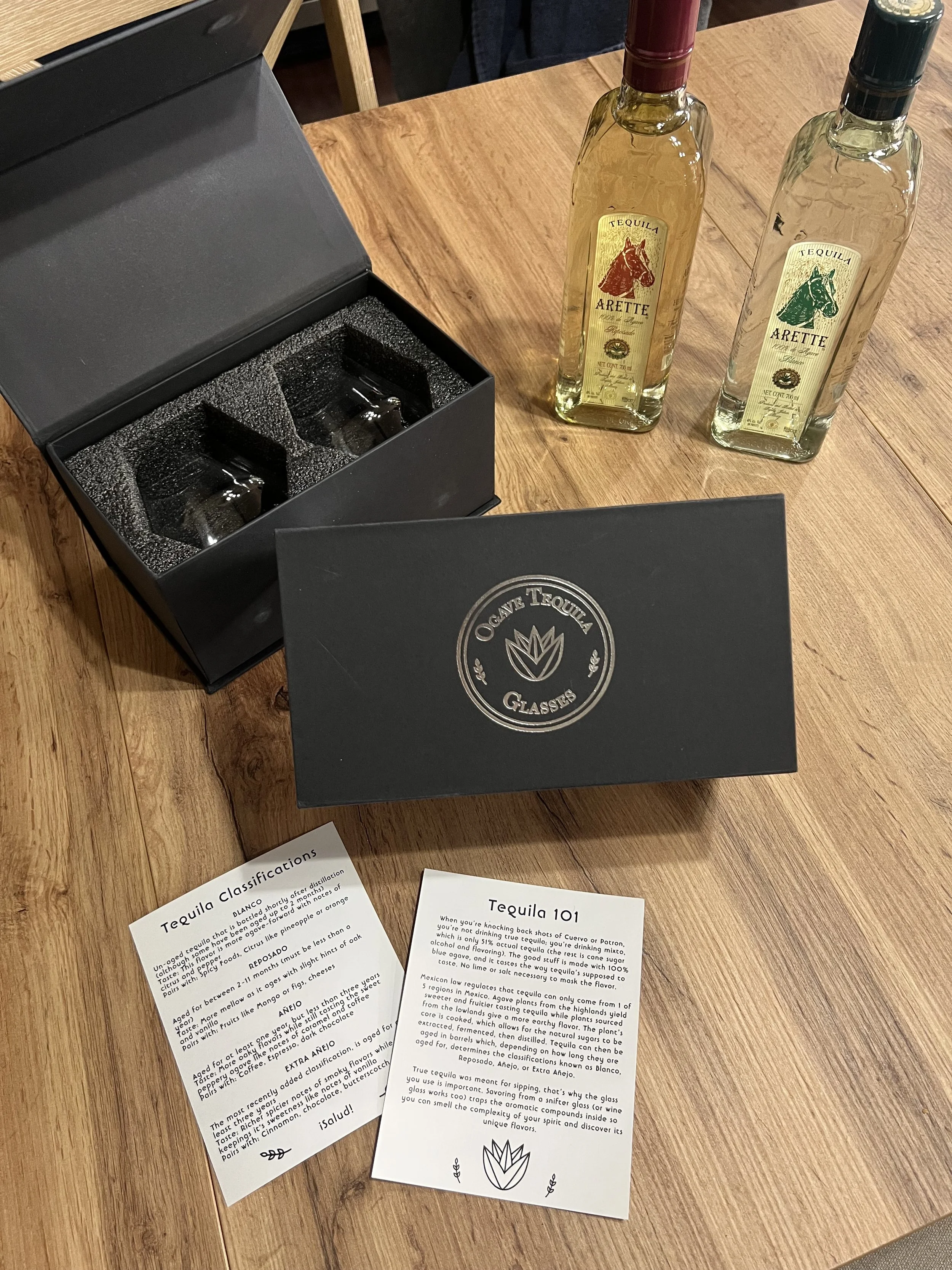 Two bottles of tequila, one with a red cap and one with a black cap, next to an open black box with glasses inside, on a wooden table, with informational pamphlets about tequila.