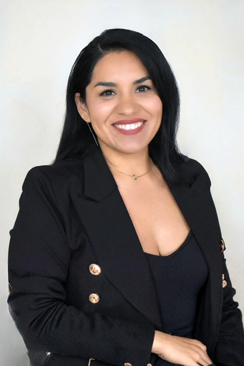 A woman with shoulder-length black hair wearing a black blazer with gold buttons, a black top, a gold necklace, and earrings, smiling against a plain light-colored background.