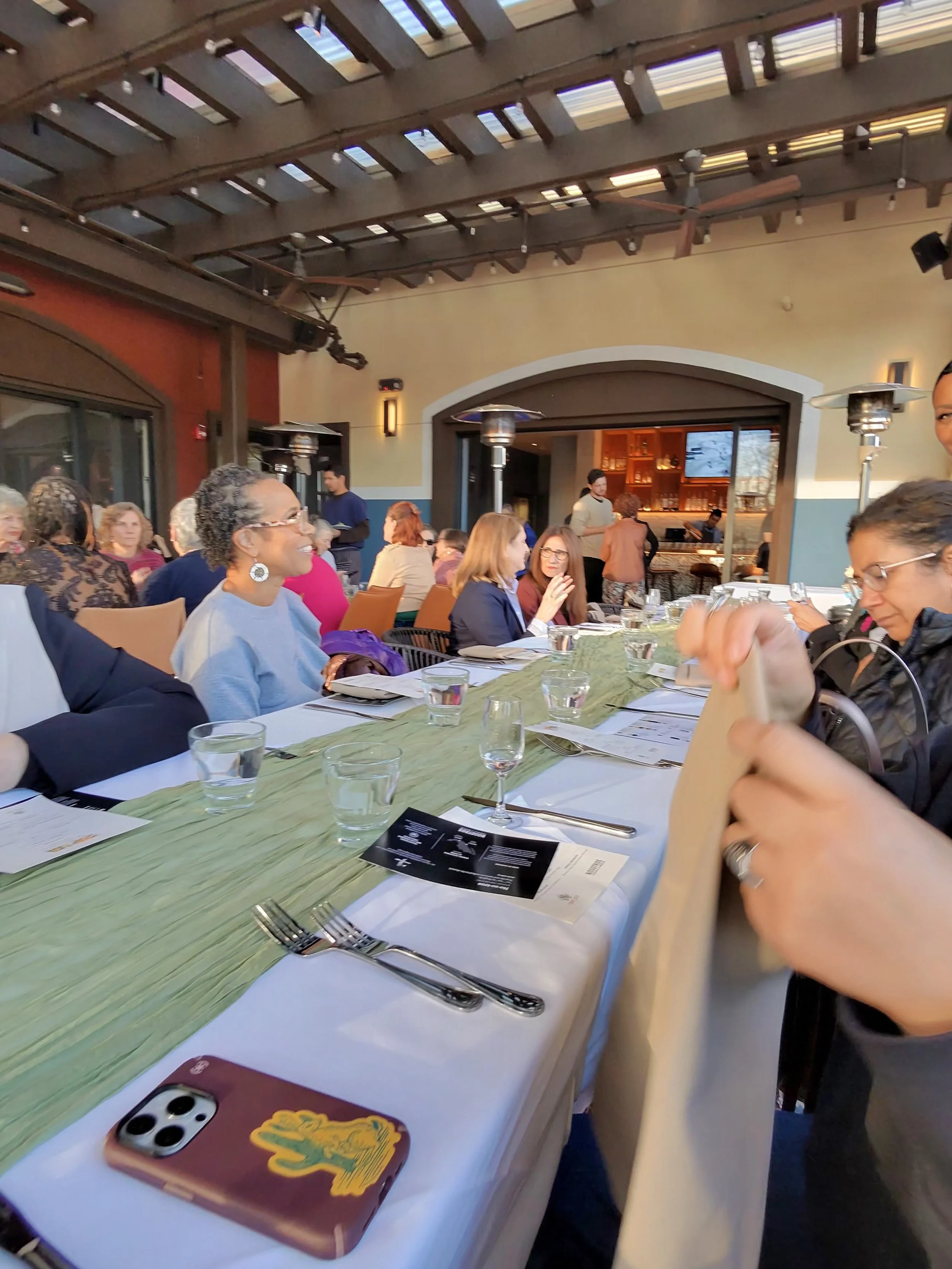 People gathered around a long dining table at a restaurant during daytime, engaged in conversation and looking at menus, with a bar area in the background and outdoor heaters visible.