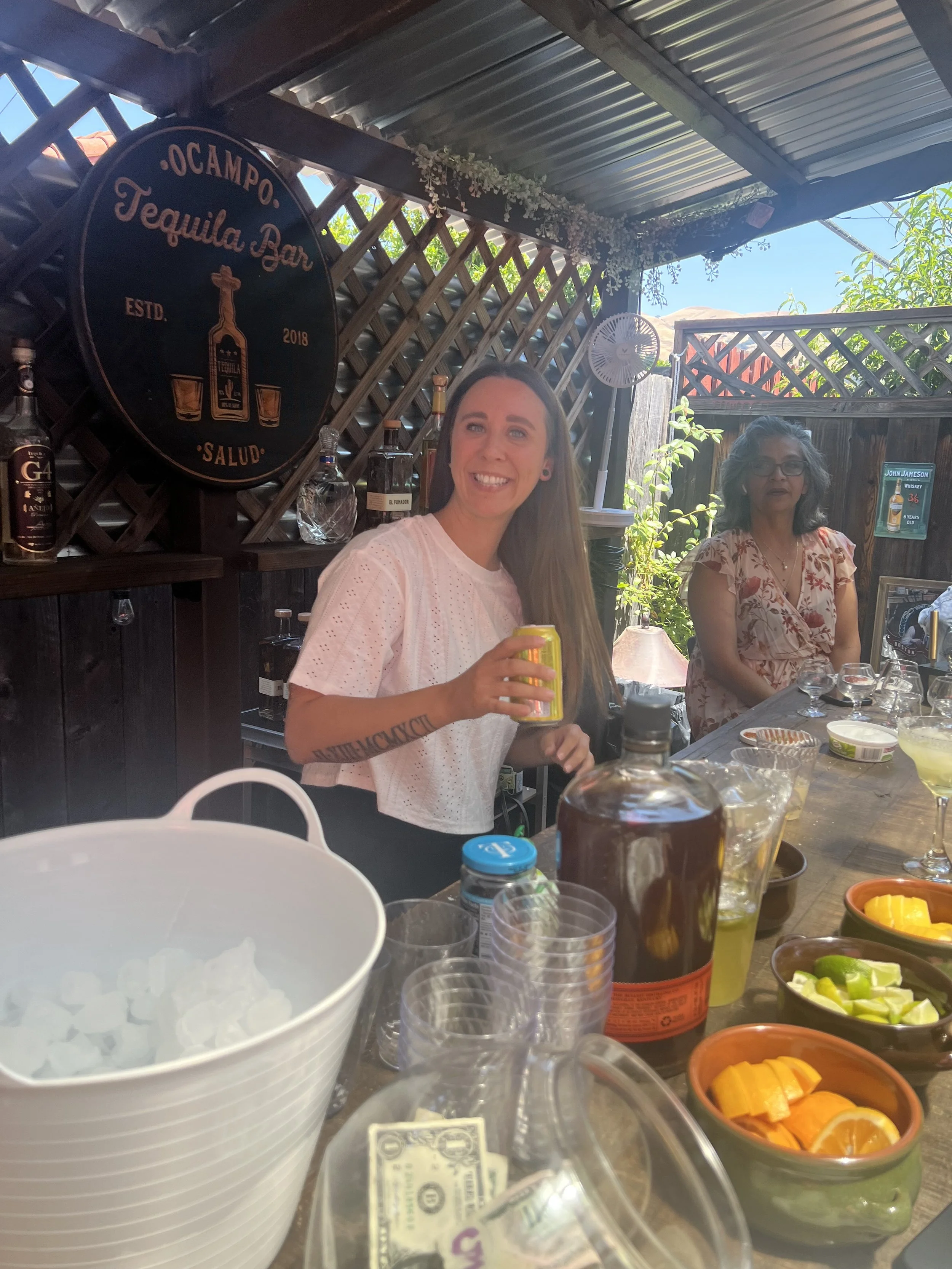 Two women at a bar with bottles of alcohol and bowls of sliced fruit on the counter, one holding a yellow can, smiling and looking at the camera, in a covered outdoor setting with a sign reading 'Ocampo Tequila Bar' in the background.