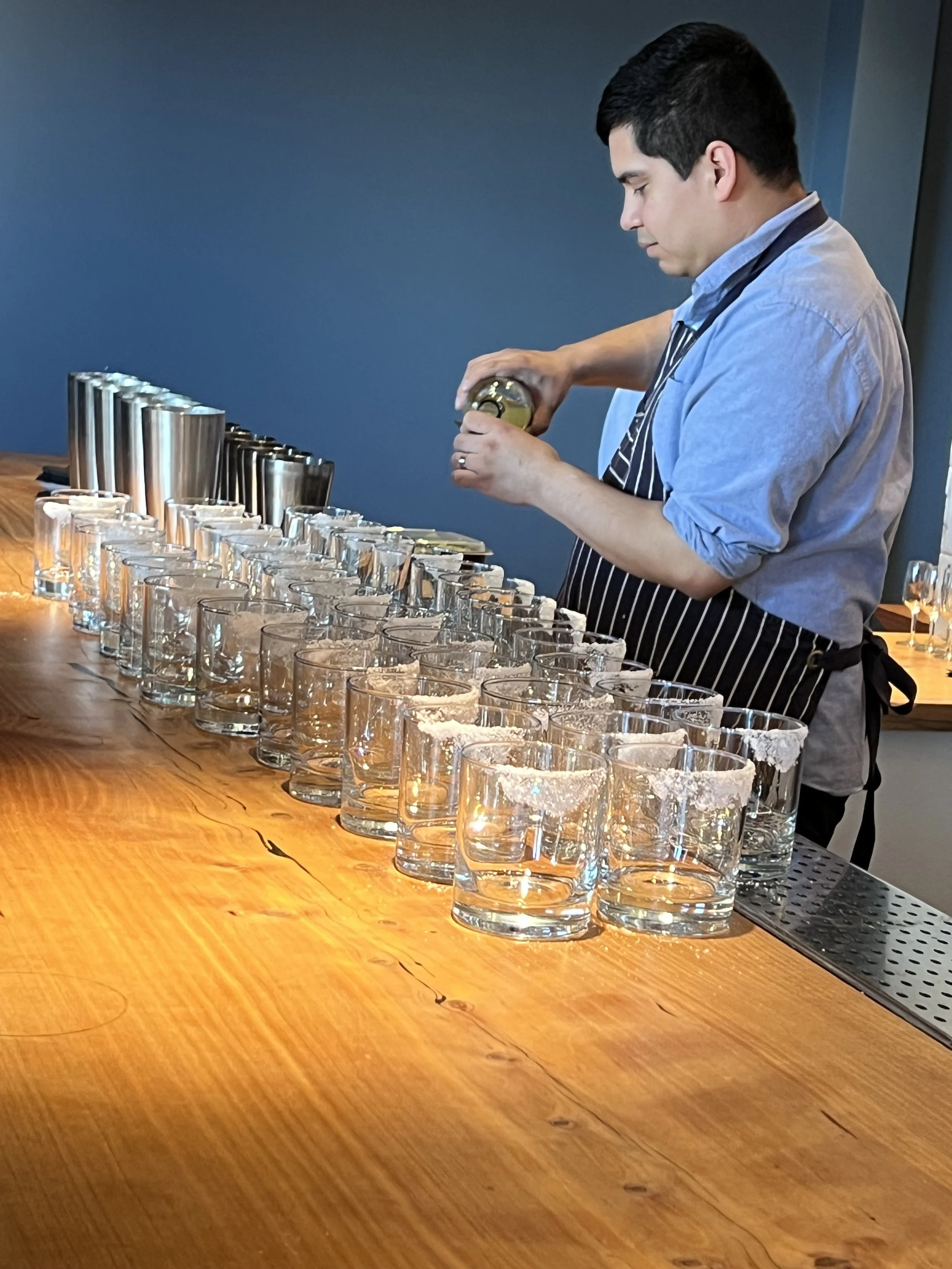 A man in a striped apron prepares glasses on a long wooden bar, with a blue wall in the background.